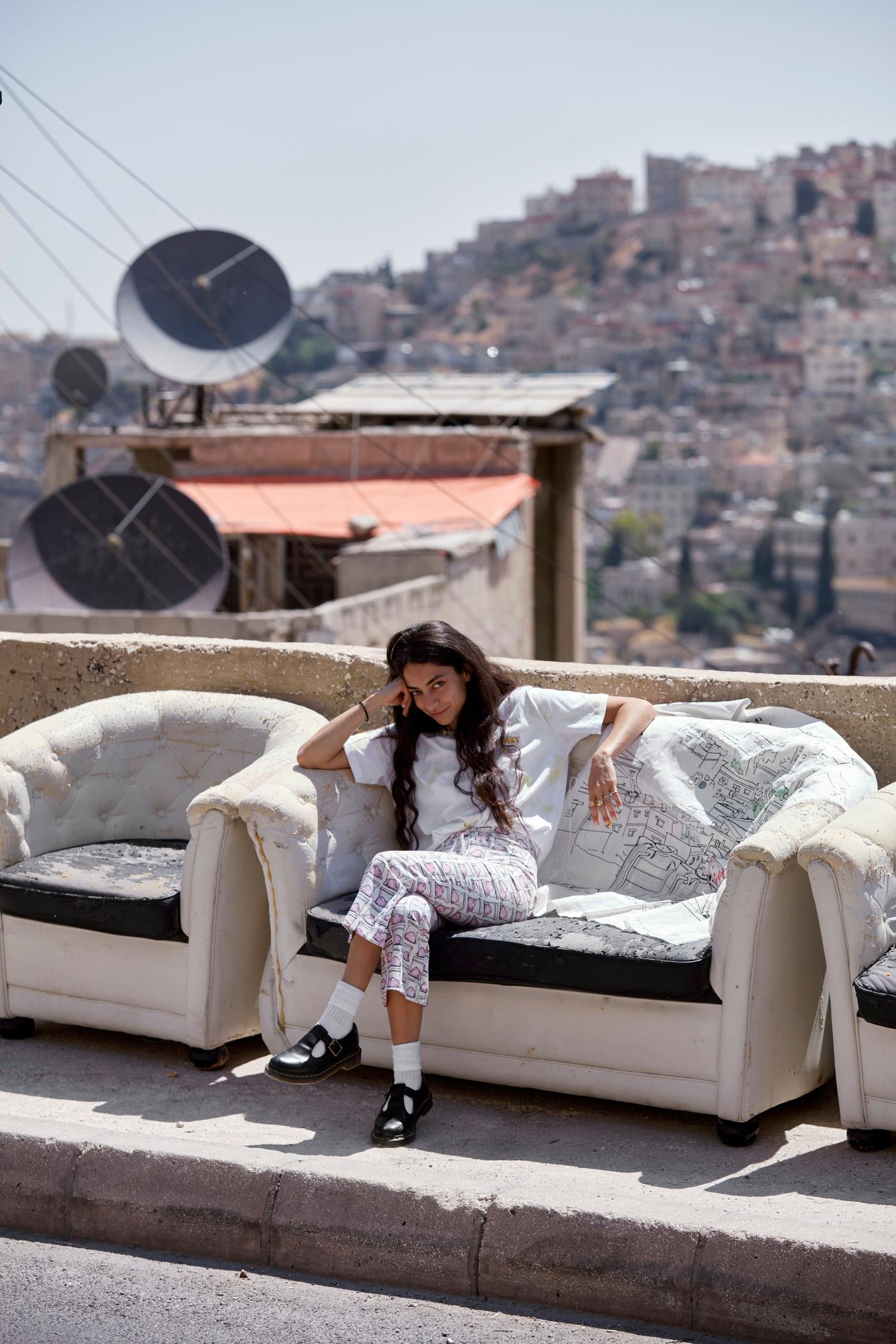 A woman sits on a sofa on a rooftop. Her head leans on her hand. She smiles.