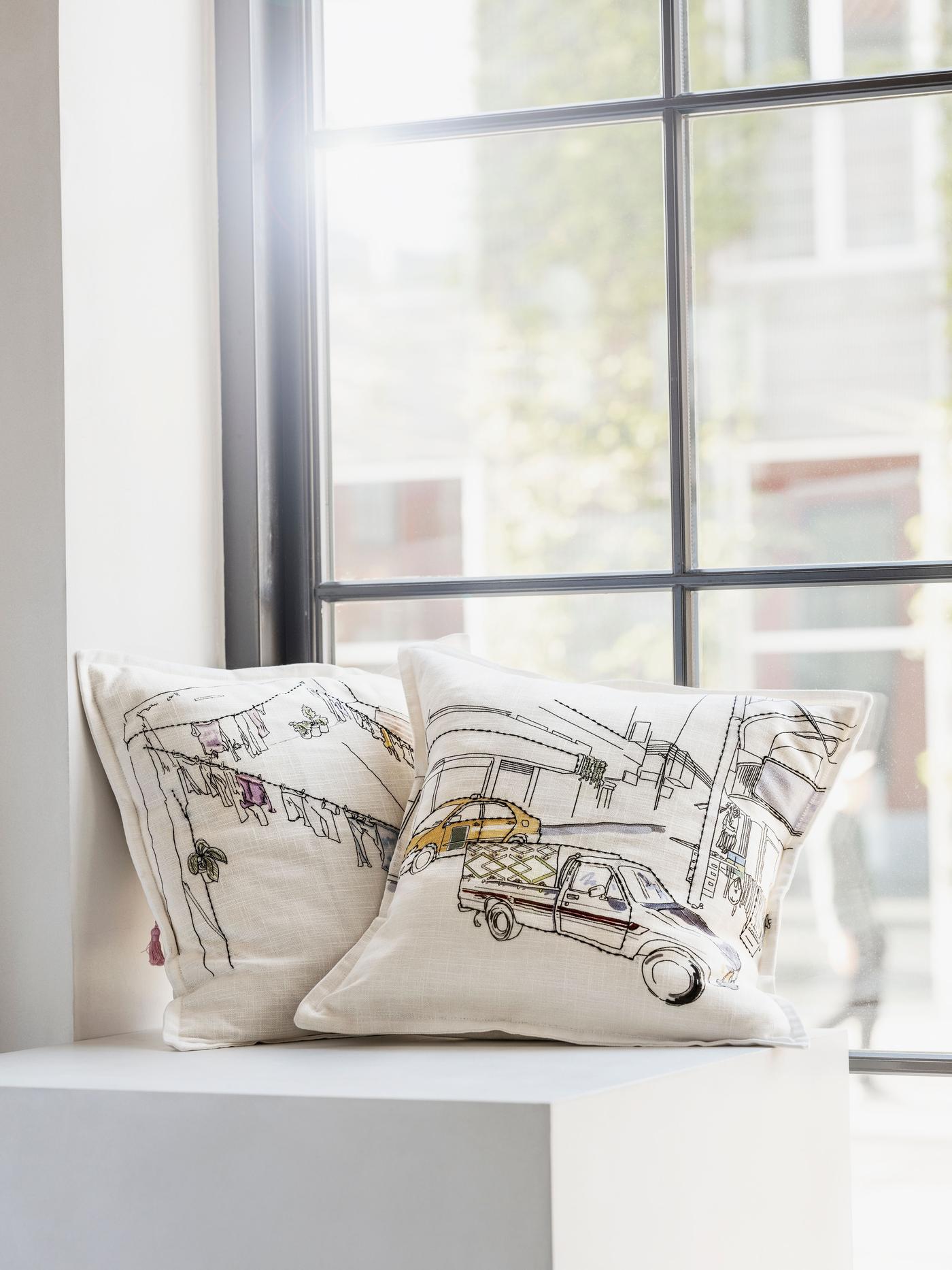 Two printed and embroidered cushions are placed on a surface, against a window.