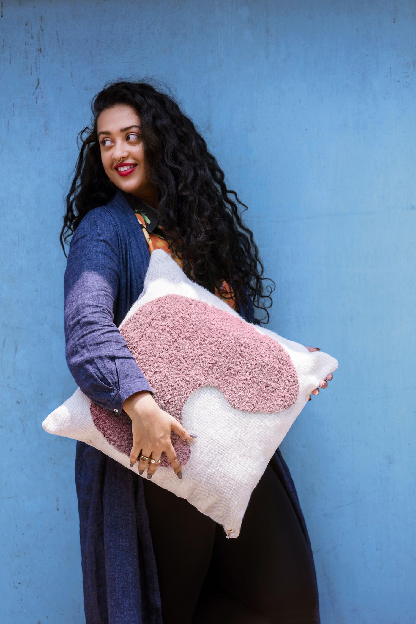 A woman smiles and stands in front of a blue background as she holds a pink and white cushion. 