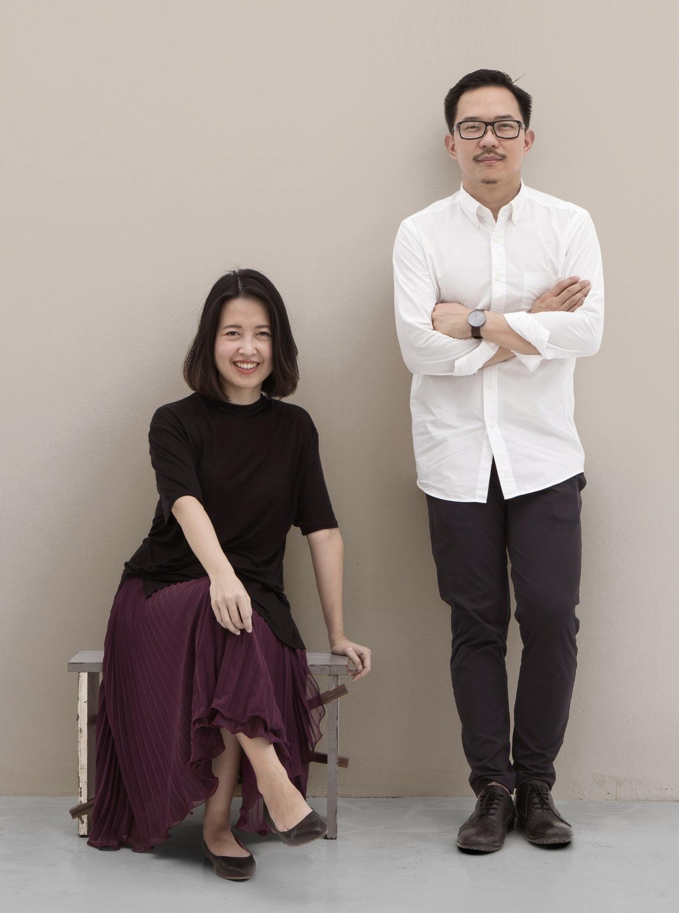 A man stands with his arms folded, while a woman sits on a chair against a beige background.