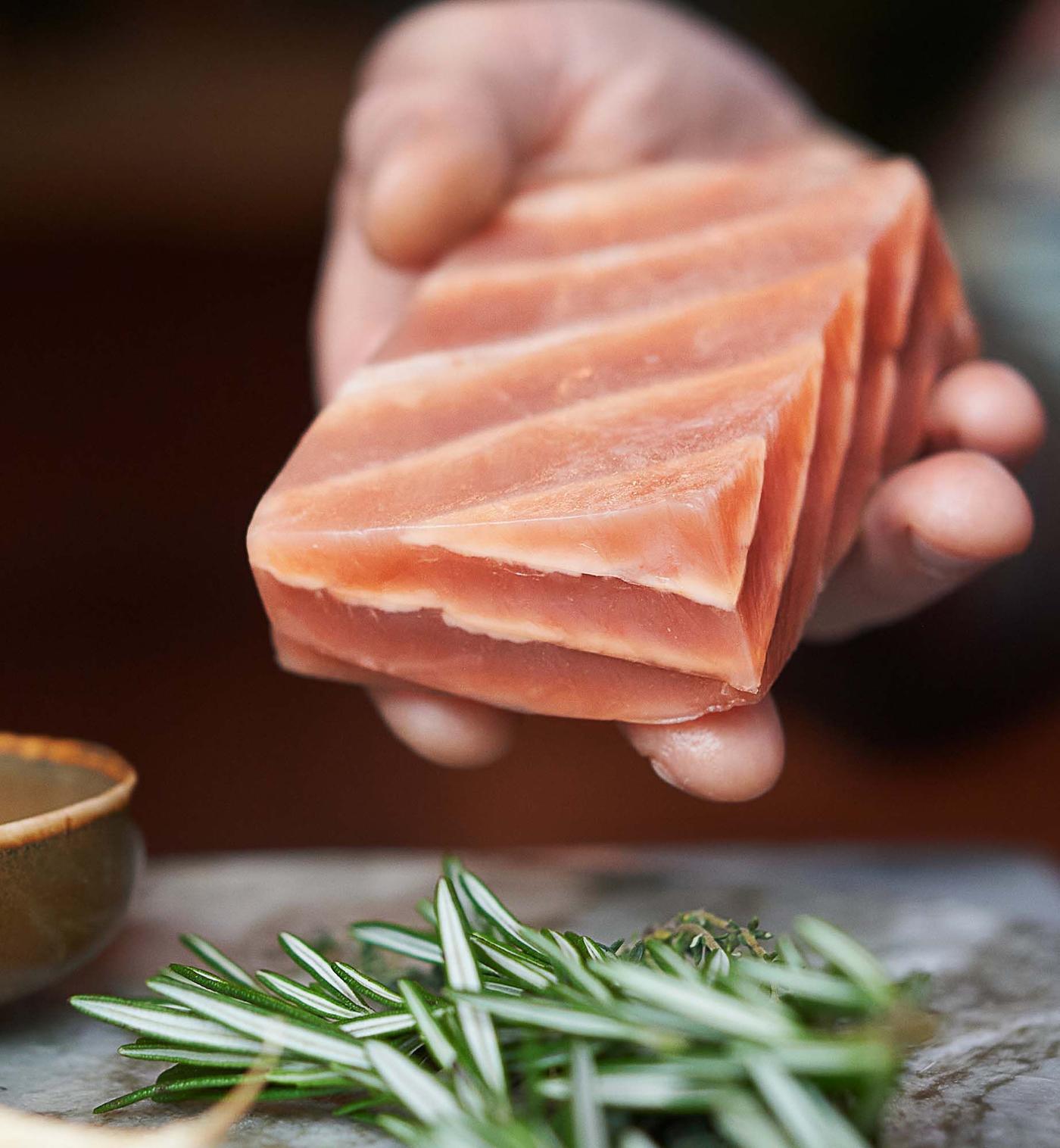 A hand holds a fillet of plant-based salmon, which looks like real salmon. Below are a few springs of rosemary.