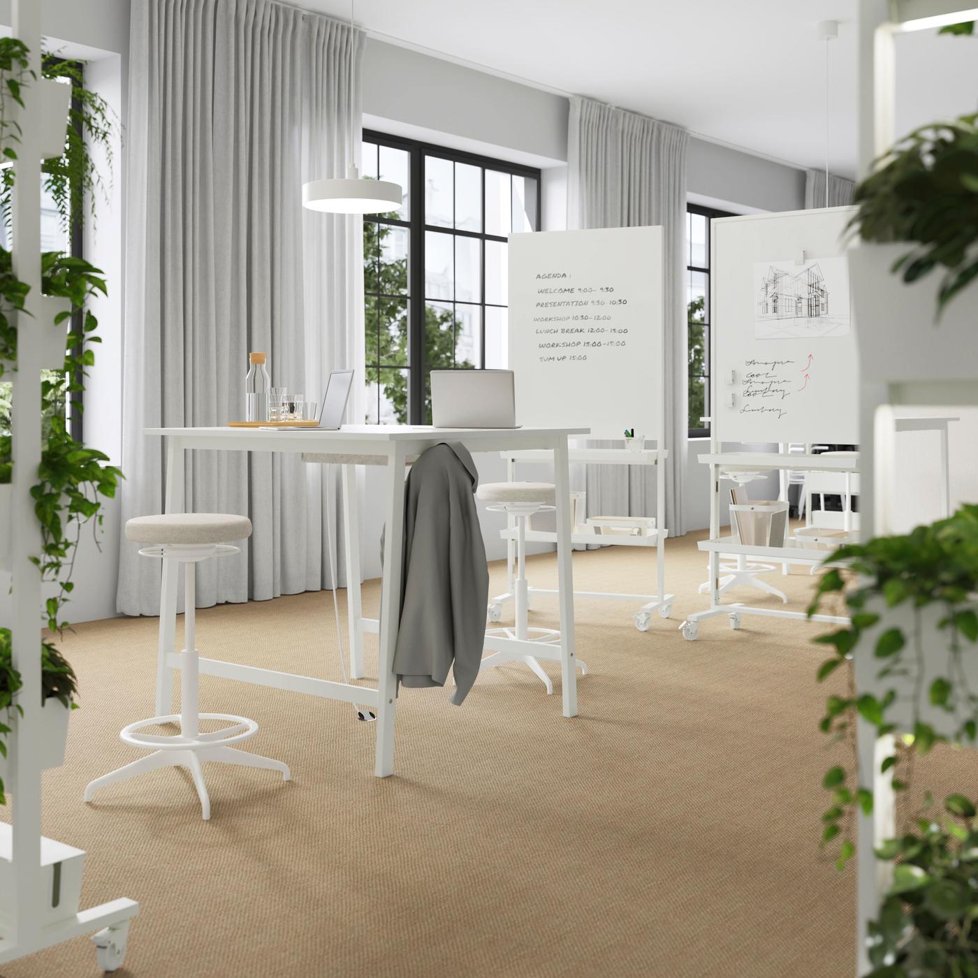 White desks, stools and a white board with text written on it. Surrounded by windows and plants.
