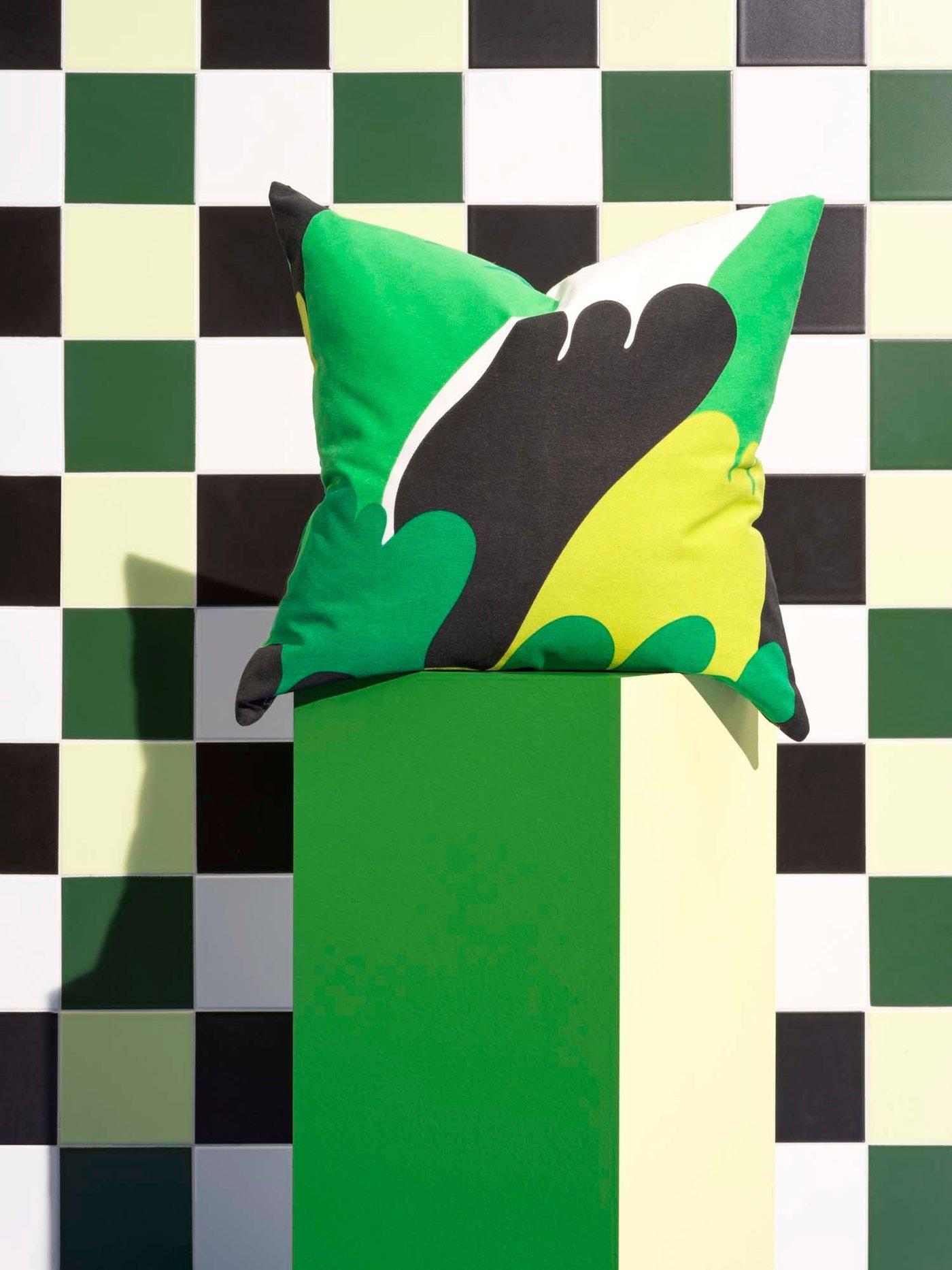 A green, black and white cushion sits on a green and yellow pedestal, against a green, white and black chequerboard background.