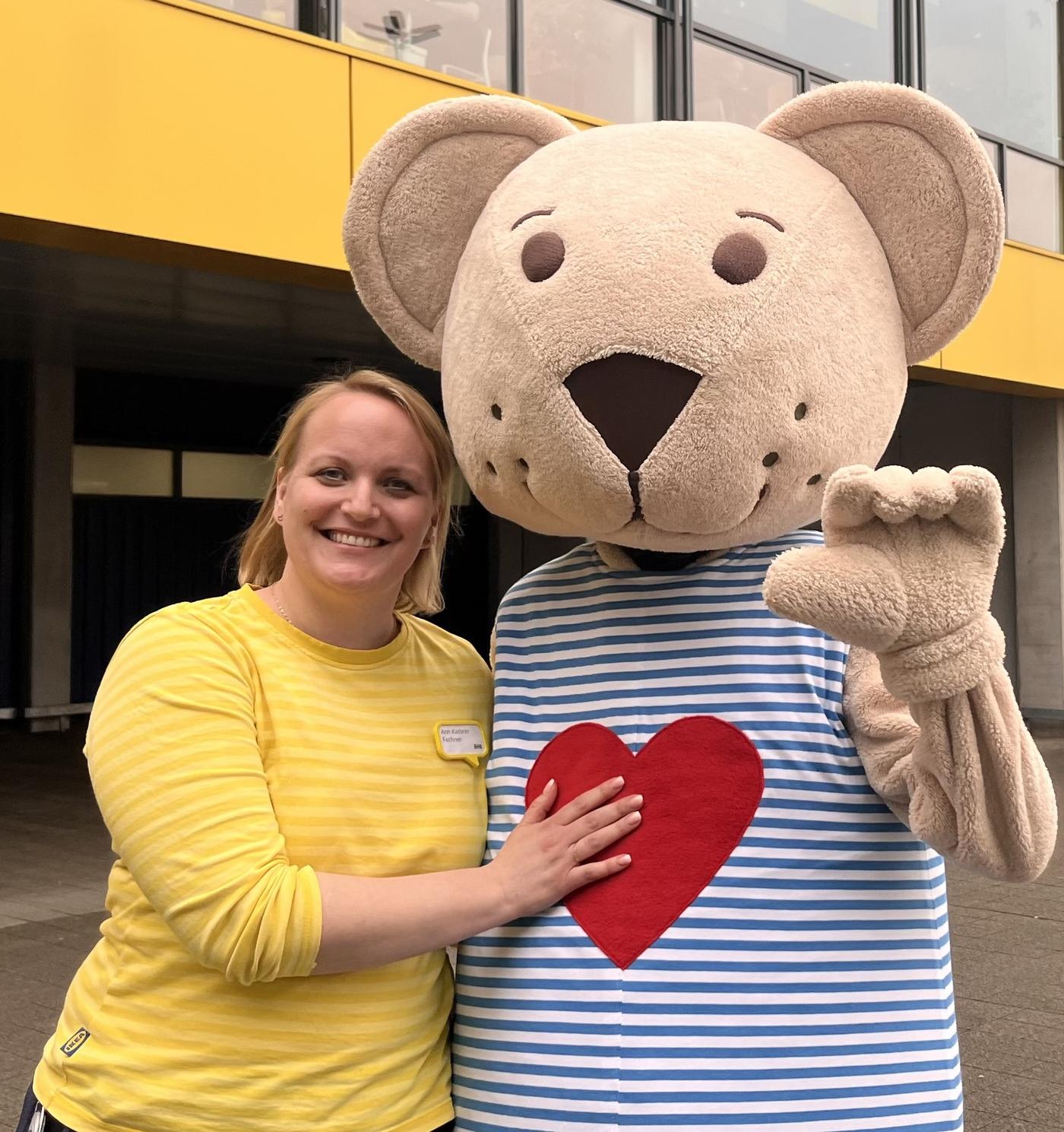 IKEA Germany co-worker Ann stands outside an IKEA store and stands next to a person in a bear costume. She smiles and touches the bear's heart.