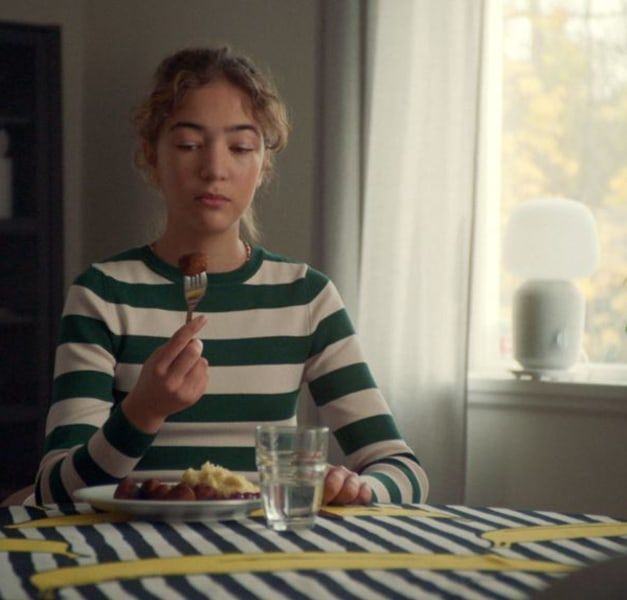 A teen in a striped top looks at the meatball on her fork. She is sitting near the window.