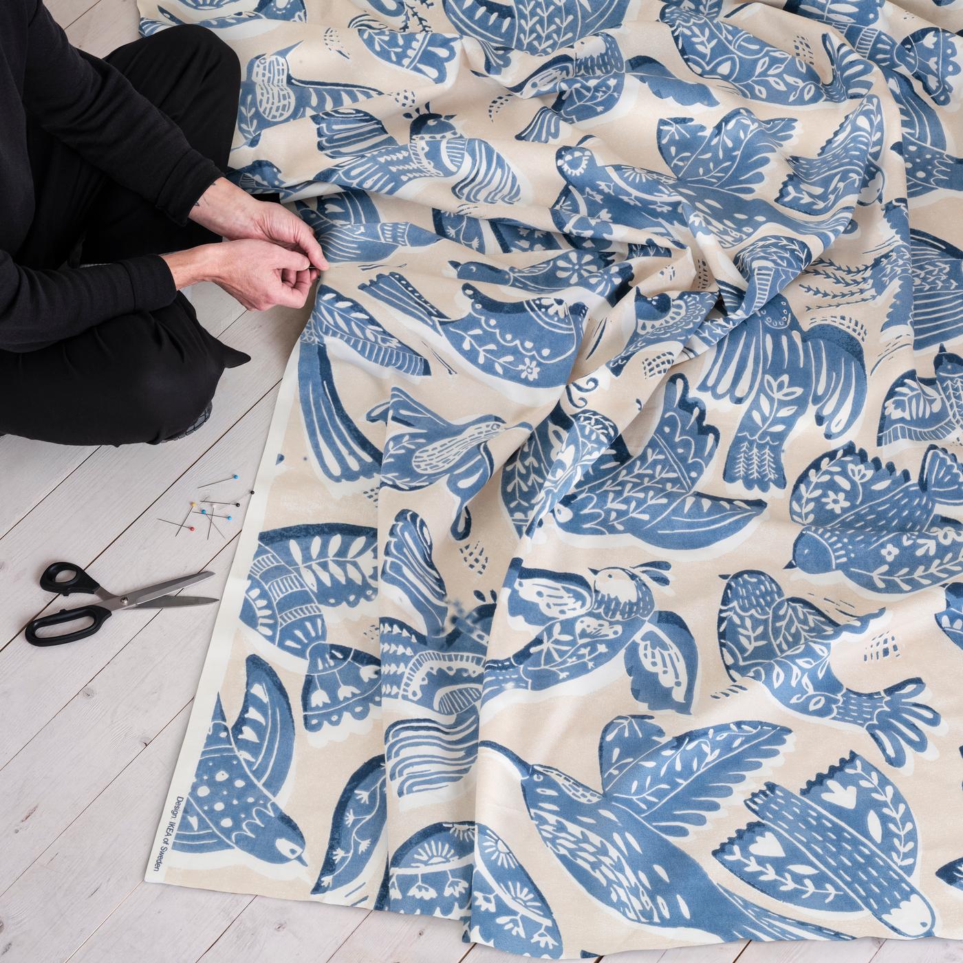 A person sits on a light wood floor, working with a large piece of BITTERSÖTTA fabric featuring a blue leaf pattern. Scissors lie nearby, indicating a craft or sewing activity.