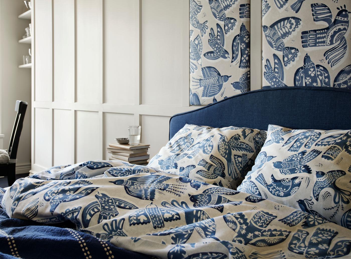 A stylish bedroom features a bed with blue and white patterned BITTERSÖTA bedding. The blue upholstered headboard complements panels of BITTERSÖTA fabric hanging above. The room exudes modern elegance with its soft colour palette and white wooden wainscoting on the walls.