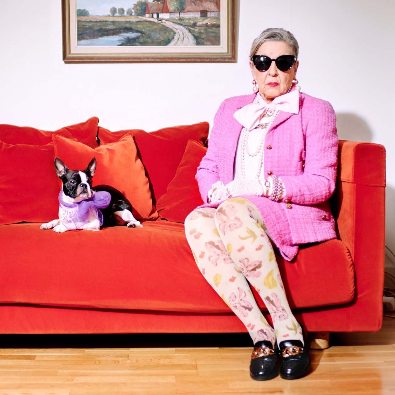 A person wearing sunglasses and a pink outfit sits in a red STOCKHOLM sofa next to a black and white dog wearing a purple bow.