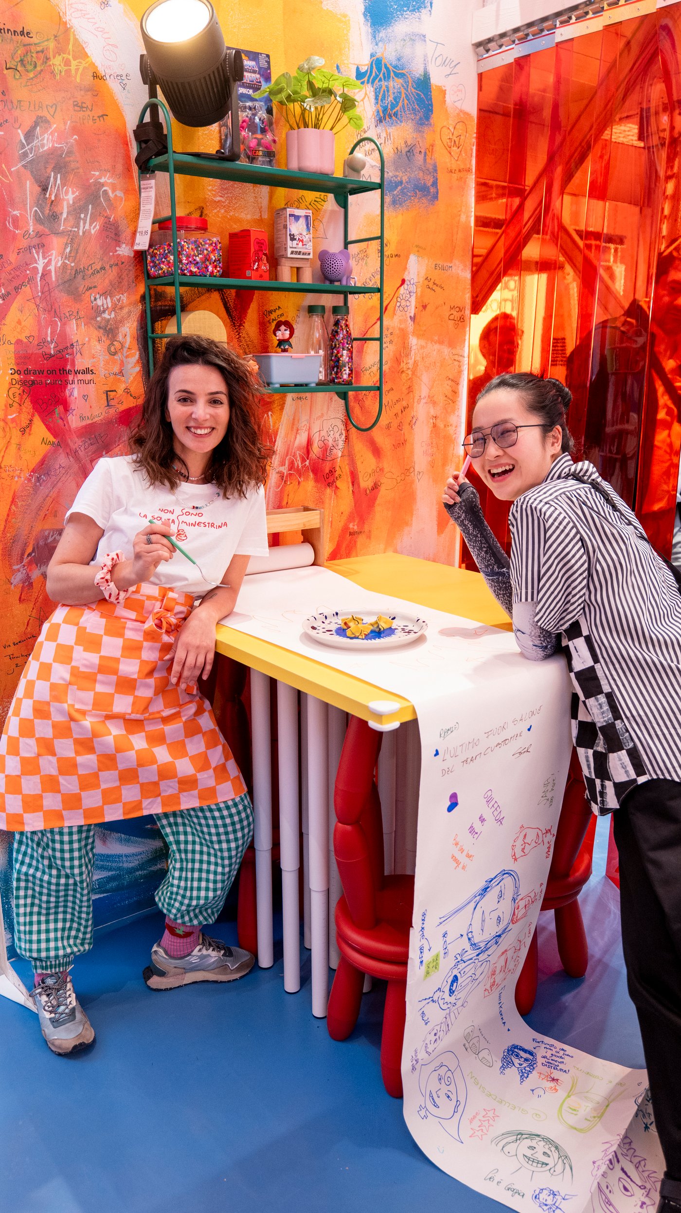 Lydia and Alessandra lean against a table in a colourful kitchen. They smile at the camera.