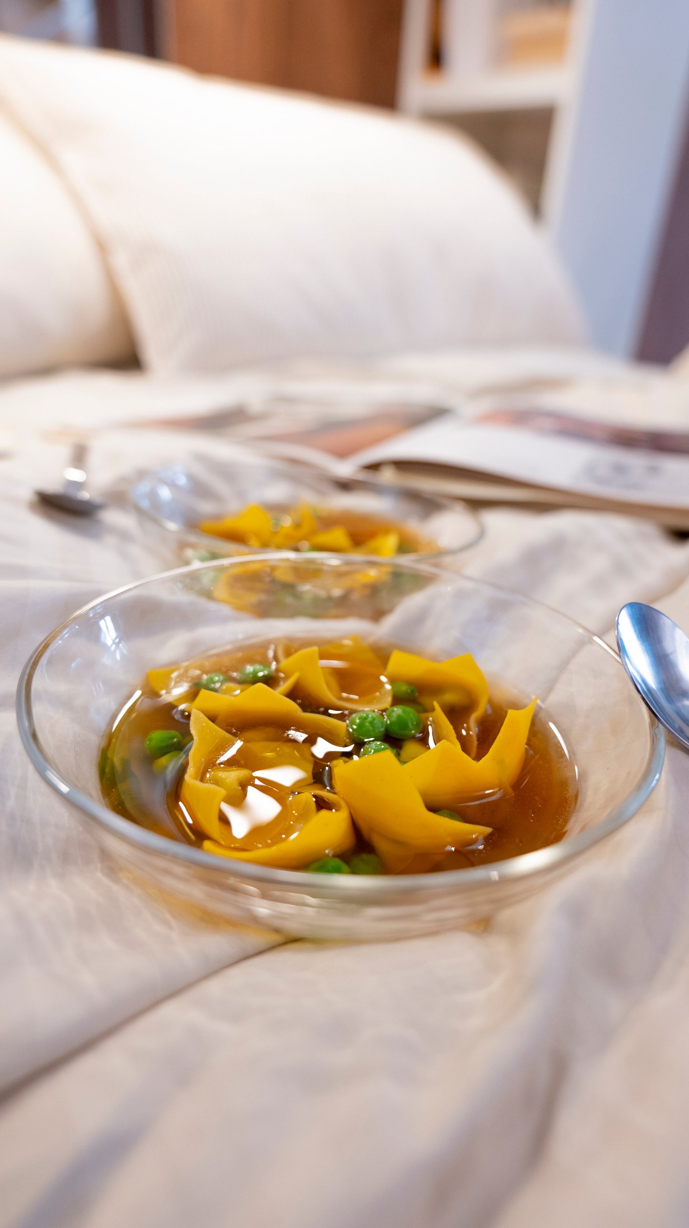 Two bowls of pasta in broth, placed on a light coloured bed.
