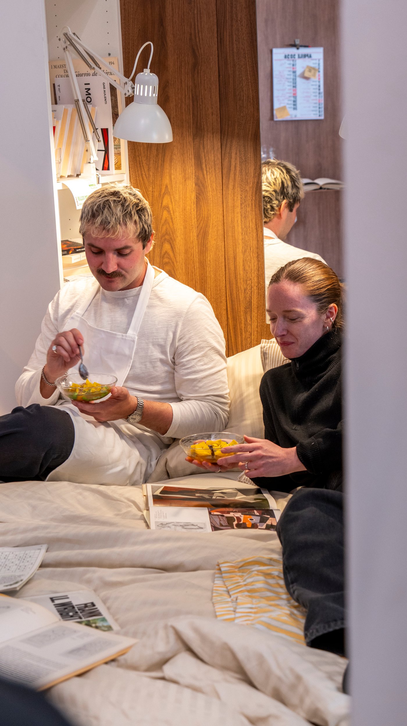 Ben and Charlotte sit on a bed in a bedroom setting. They are eating pasta in broth, while looking at a magazine.