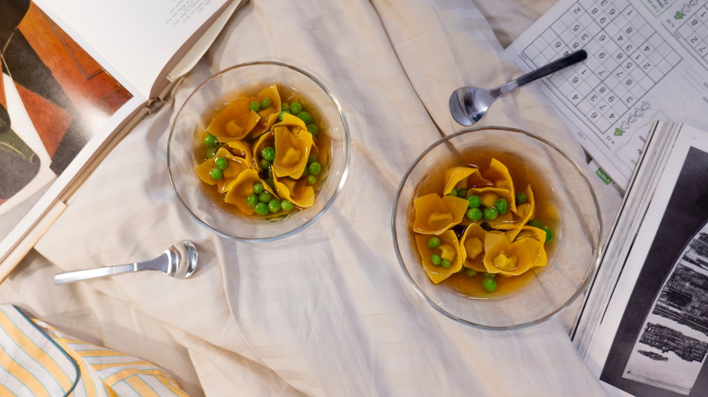 An aerial view of two bowls of pasta in broth on a bed, next to drawings and books.