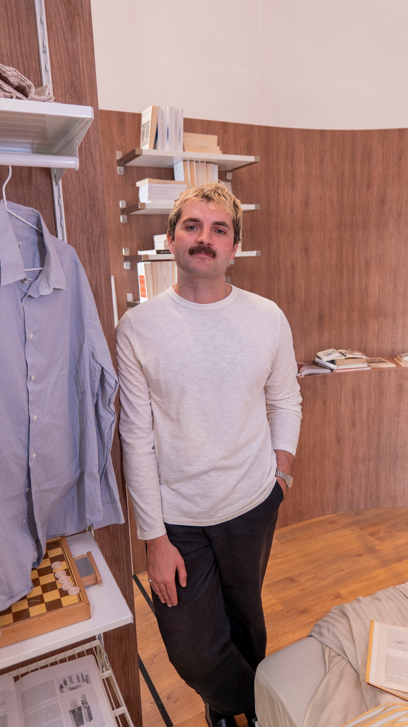 Ben Lippett leans against a wall, near a shirt hanging on a rail in a bedroom setting.