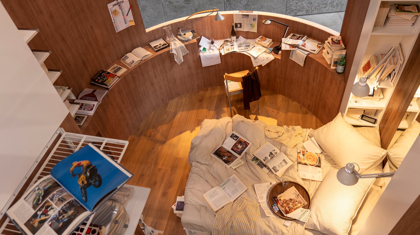 An aerial view of a messy-looking curved bedroom, with papers and open magazines strewn everywhere.
