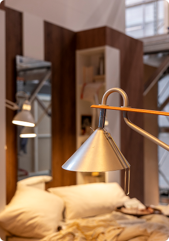 An illuminated metal lamp, with a messy bed in the background.