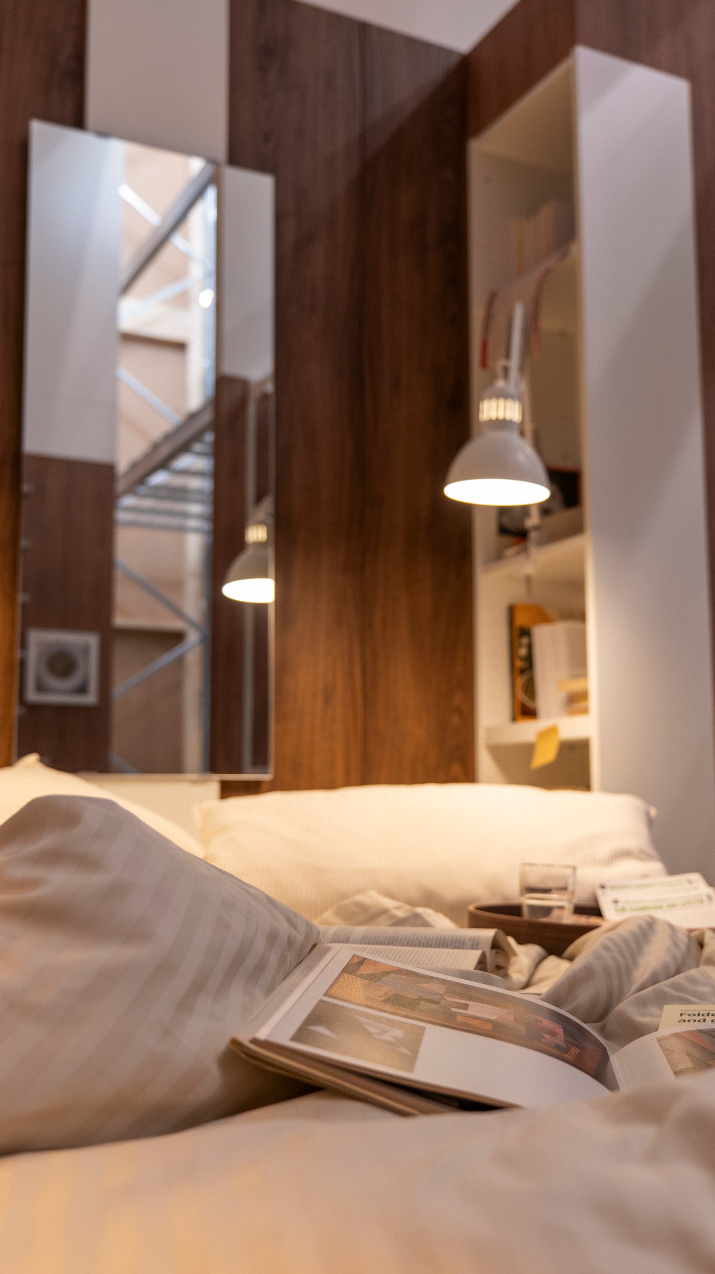 A bedroom with an illuminated lamp in the background and bed, attached to a storage unit.