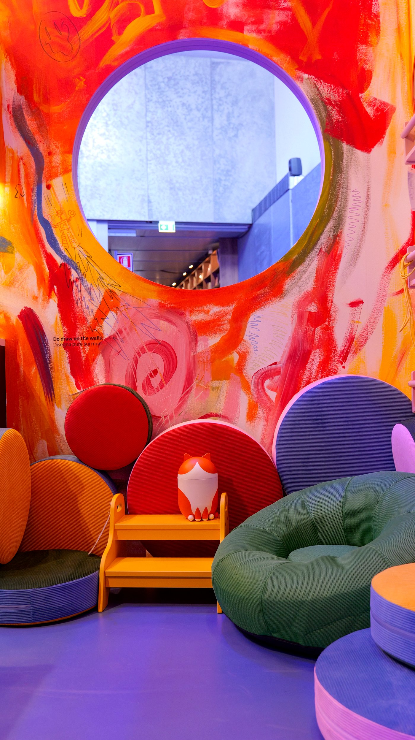 A large, round mirror on a colourful walk above six GREJSIMOJS floor cushions and a cat-shaped GREJSIMOJS storage with lid.