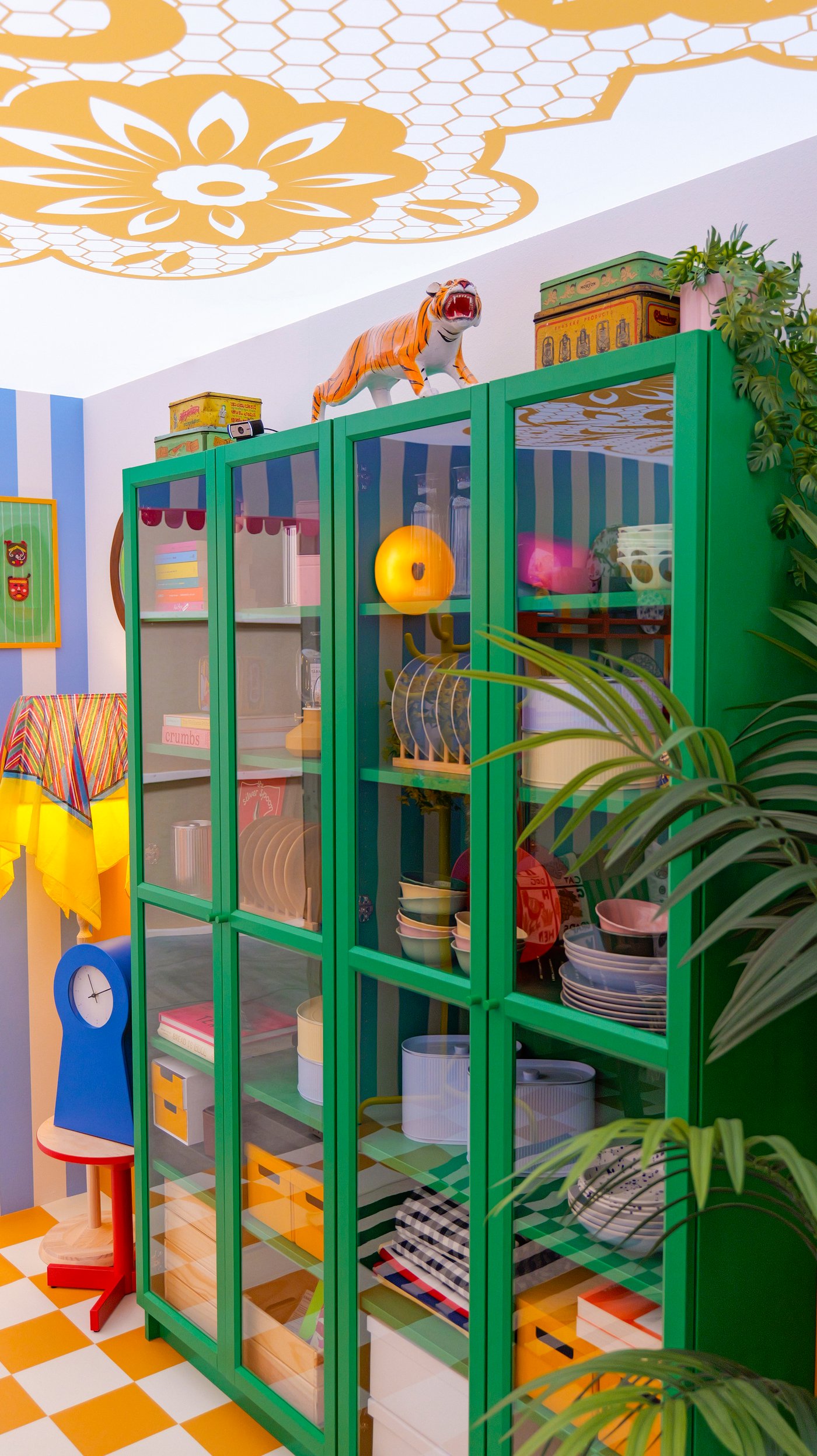 A green glass-fronted cabinet filled with colourful dinnerware, with novelty objects on top.