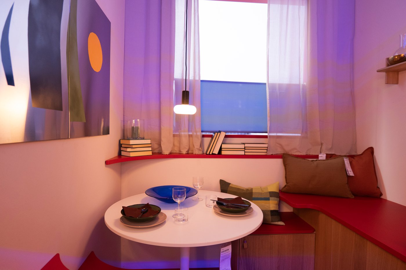 A dining-bed area in a studio, with books on the windowsill and a lamp over the table.