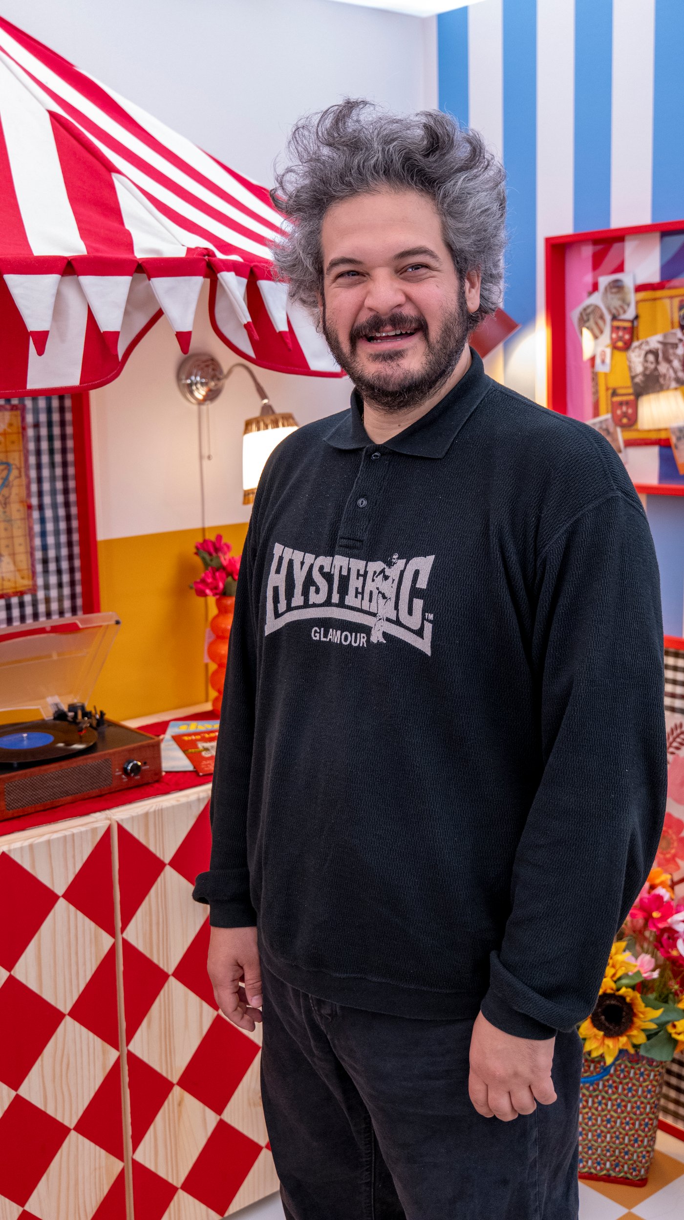 Maurizio smiles at the camera. He is standing next to a red and white diamond pattern table with a matching striped awning.