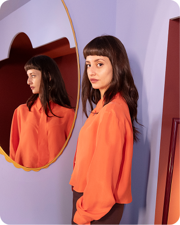 Maye stands next to a mirror on a lilac wall with her reflection. She looks at the camera.
