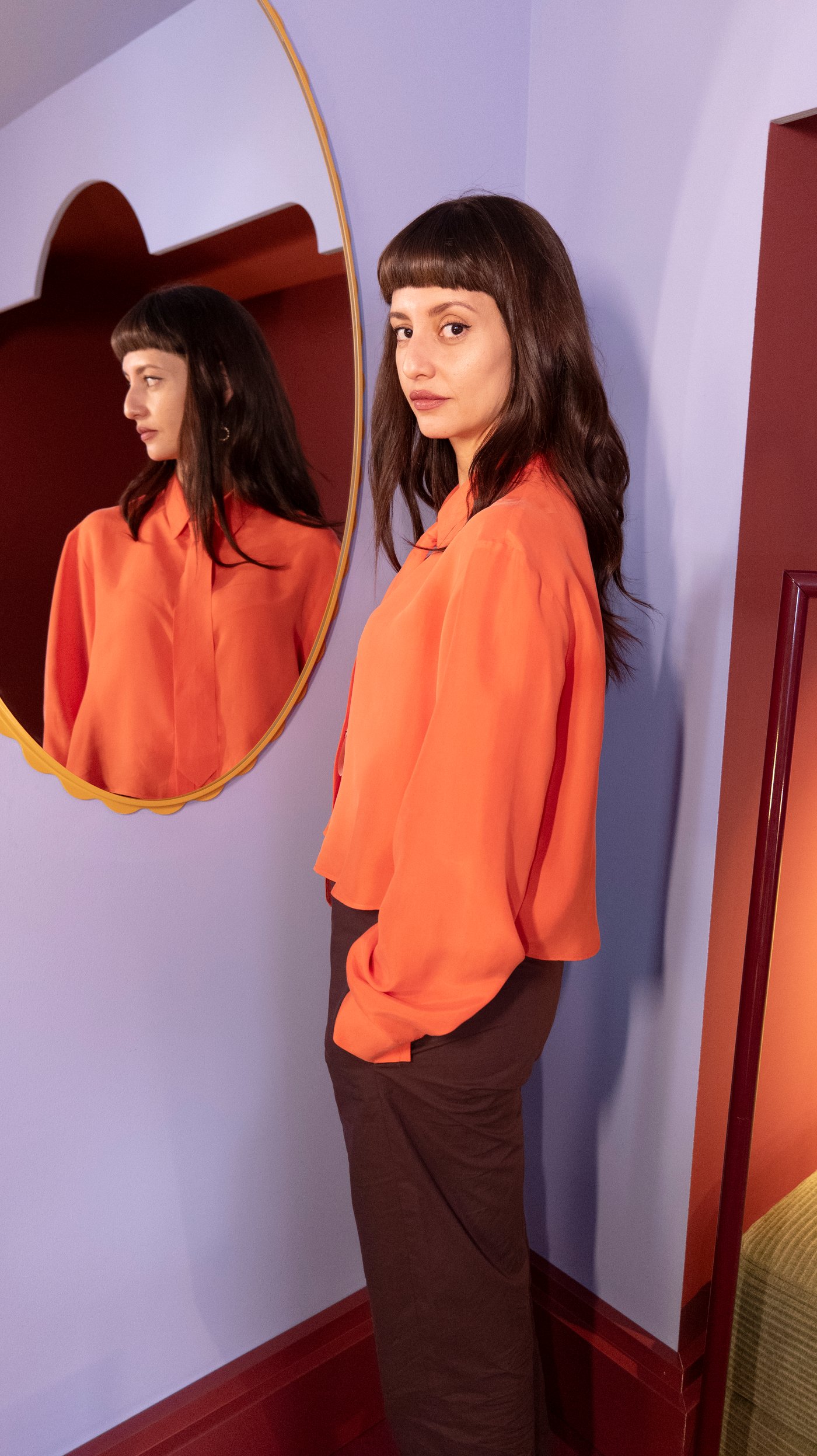 Maye stands next to a mirror on a lilac wall with her reflection. She looks at the camera.