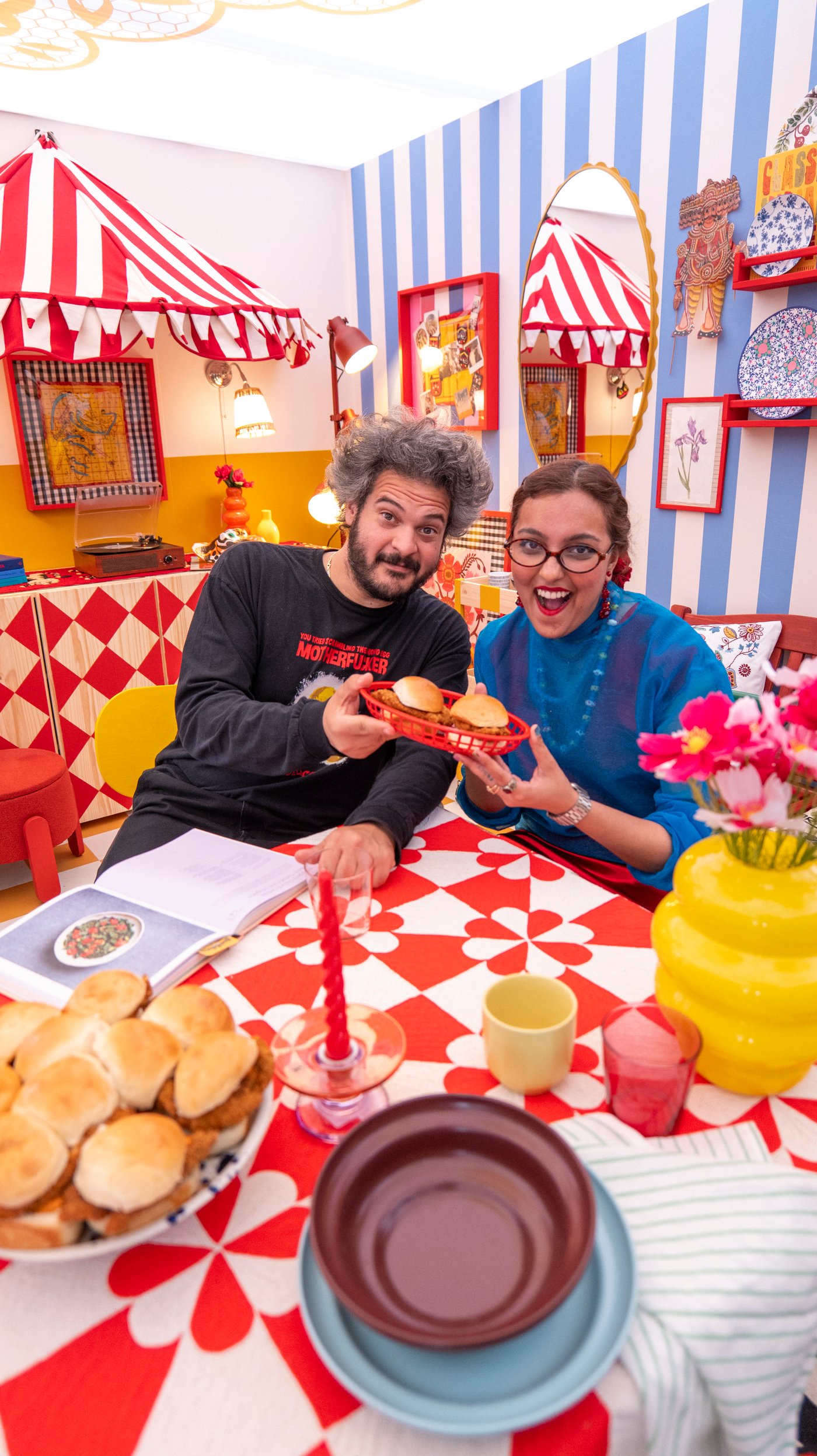 Two people sit at a table in a graphic, brightly coloured dining room at a table. They smile at the camera while holding a basket of burgers..