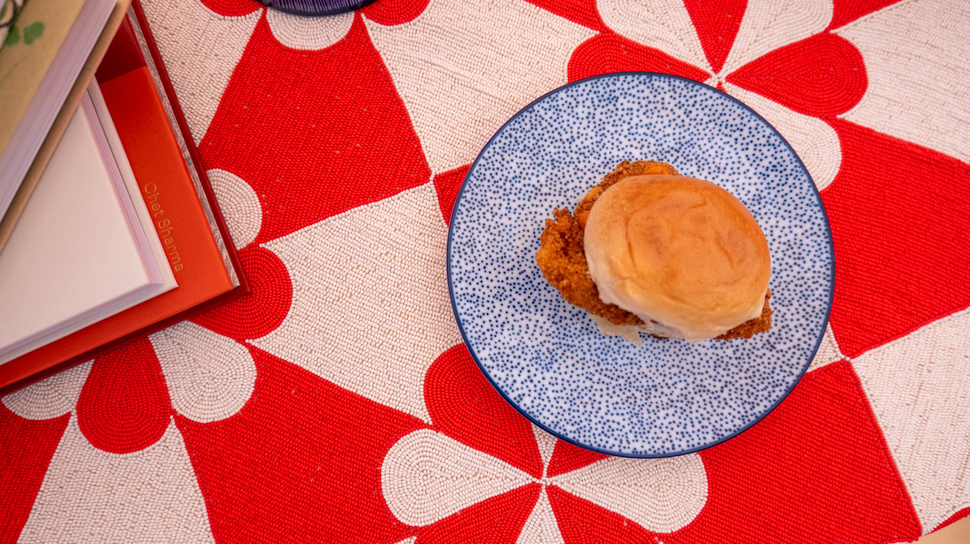 An aerial view of a chicken burger on a blue and white plate, against a red and white graphic tablecloth.