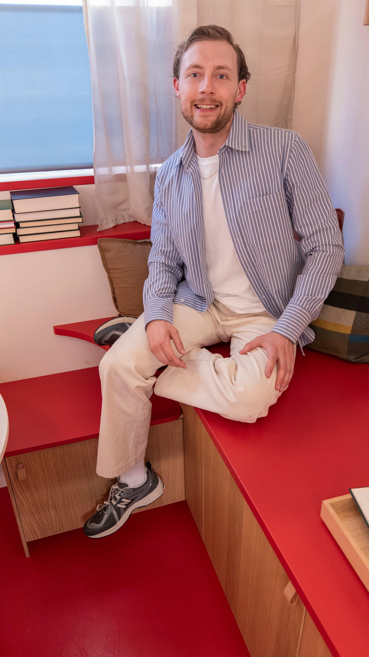 Oliver sits on a red bench-bed. He smiles at the camera.