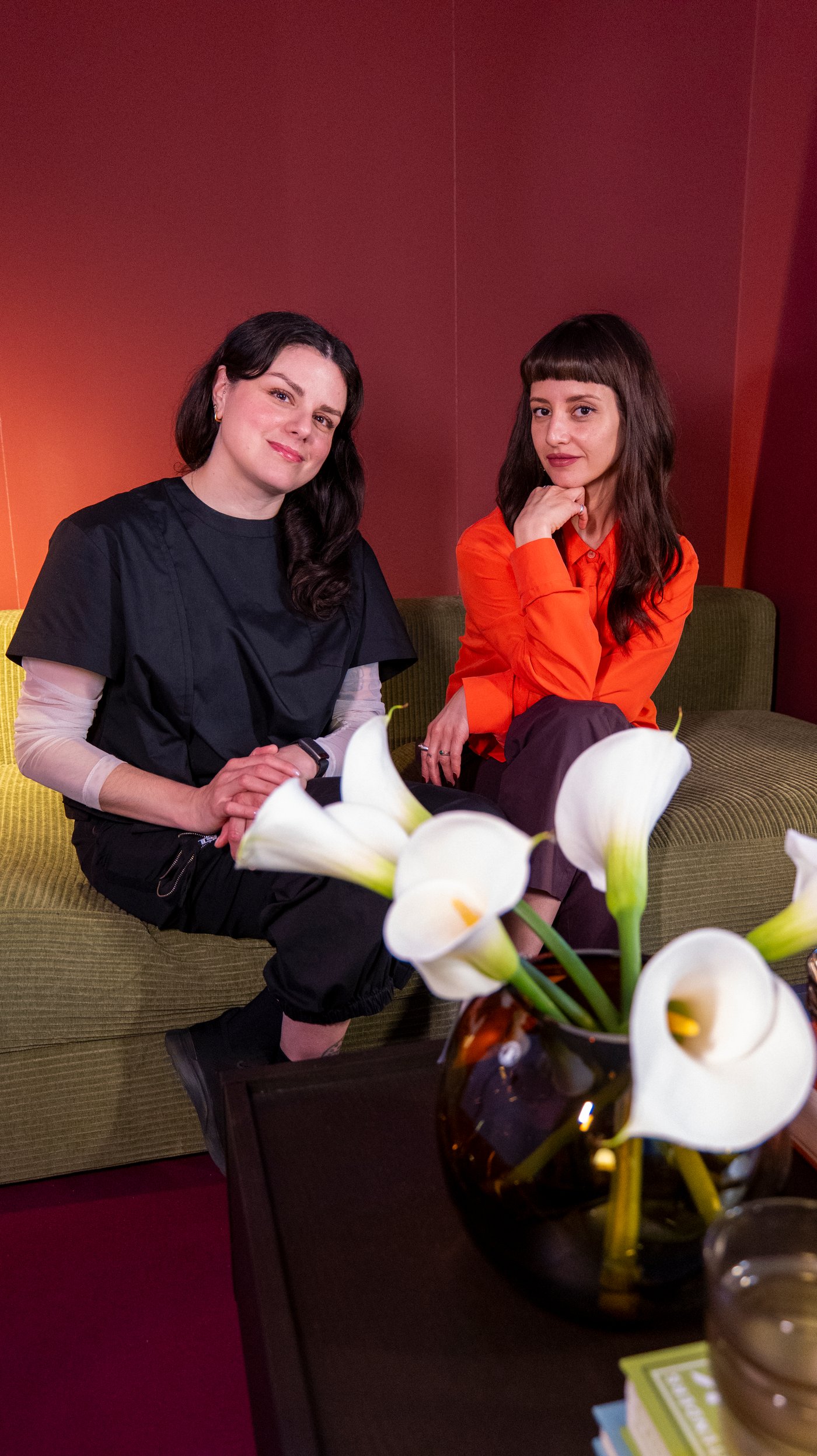 Rosio and Maye sit on a green sofa in a dark red room, with lillies in the foreground. They look at the camera.