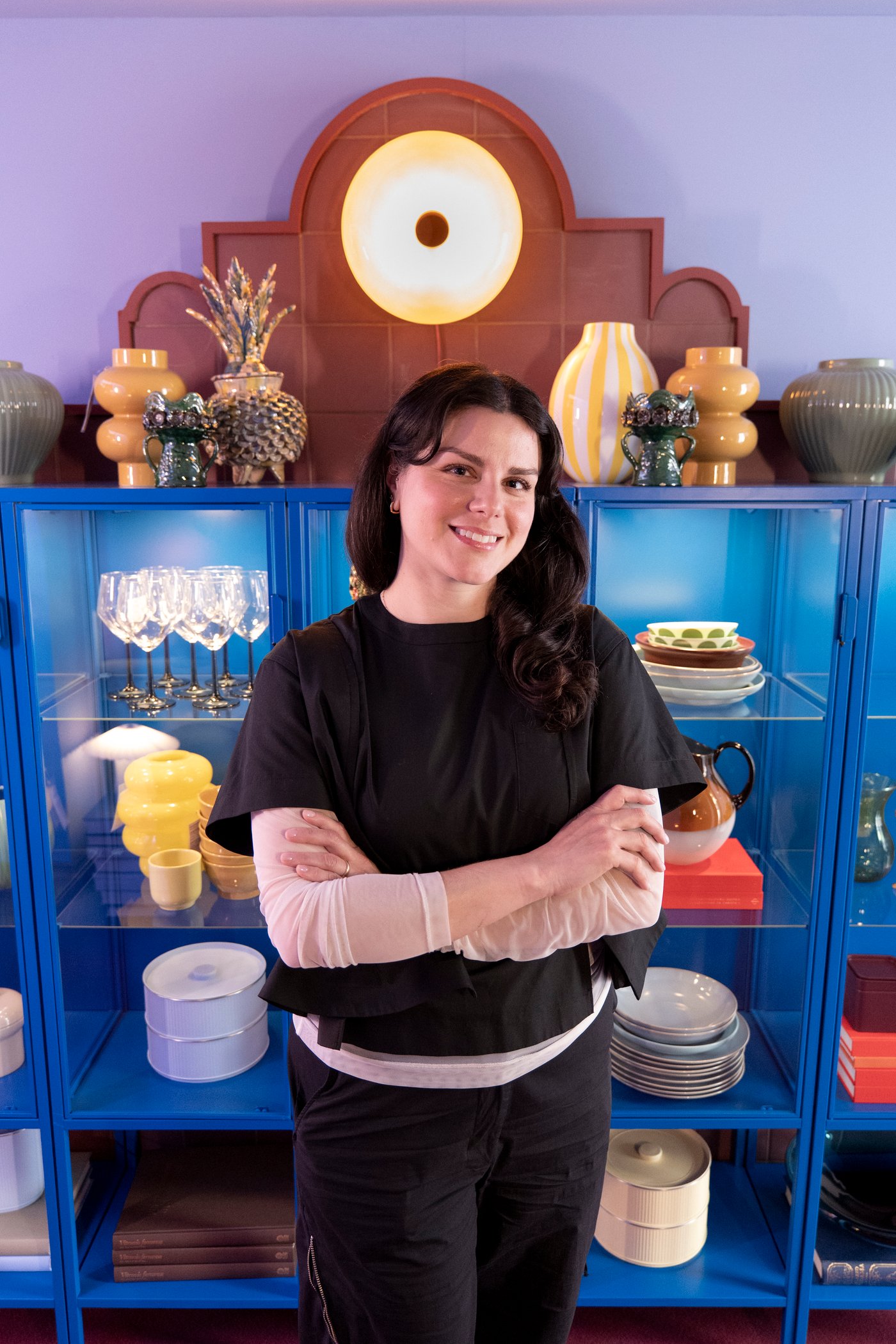 Rosio stands in front of a blue, glass-front storage unit filled with plates and glasses. She smiles at the camera.