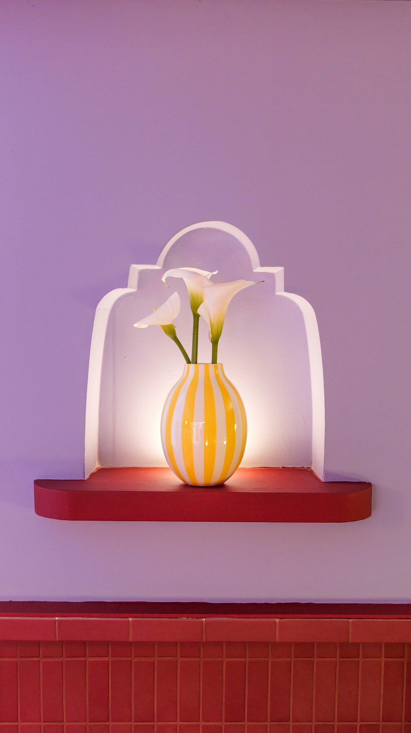 A red, backlit shelf on a lilac wall, with a yellow and white striped vase.