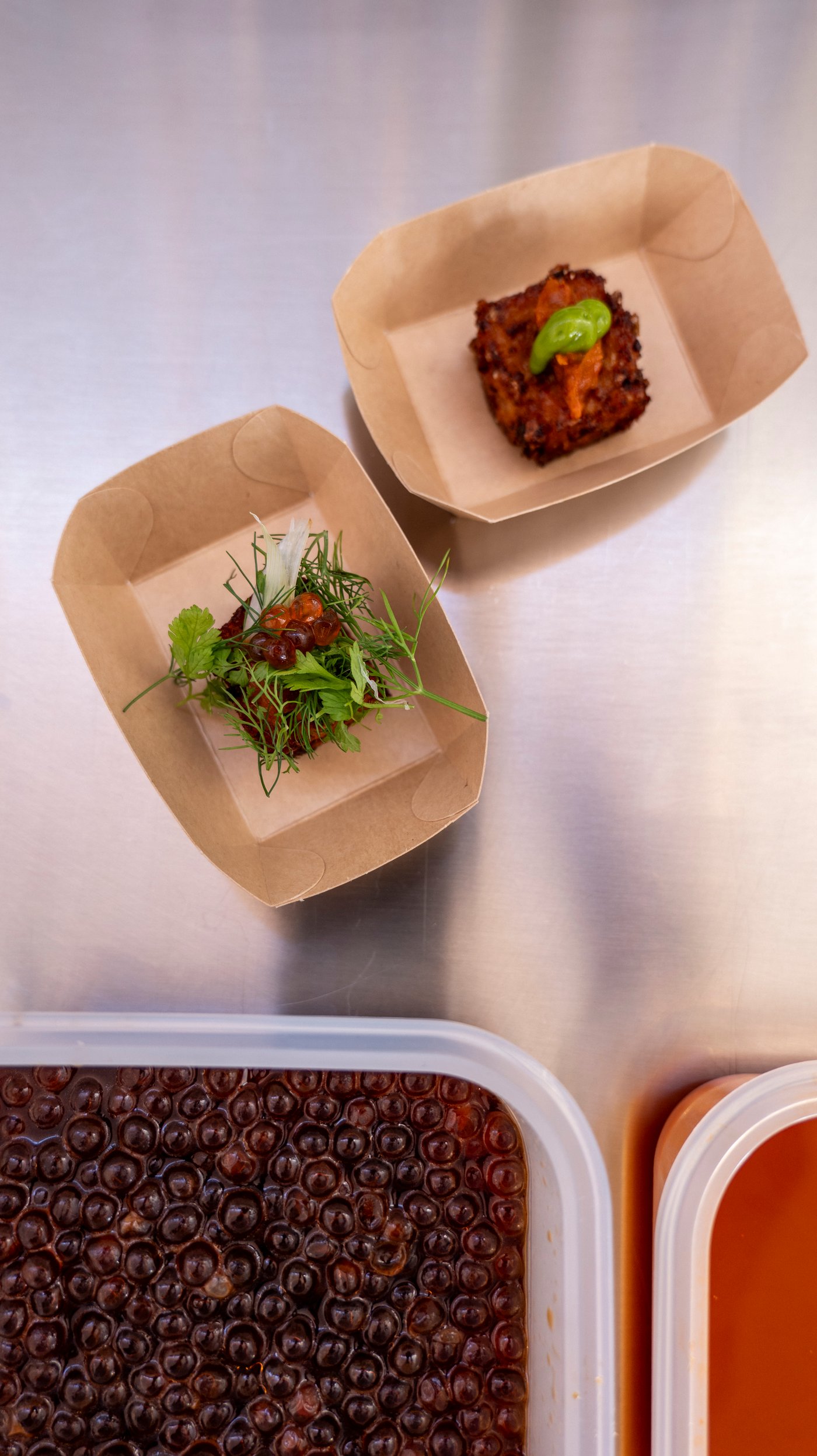 An aerial view of two kimchi rice bites in recyclable takeaway packaging on a metal surface,