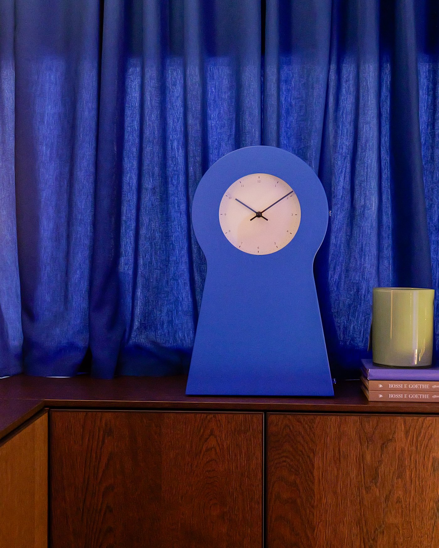 A blue clock on a wooden surface in front of a blue curtain.