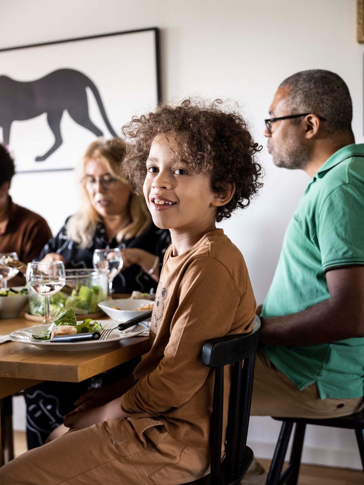 The family sit at the dining table enjoying lunch. Victor is looking at the camera and smiling.