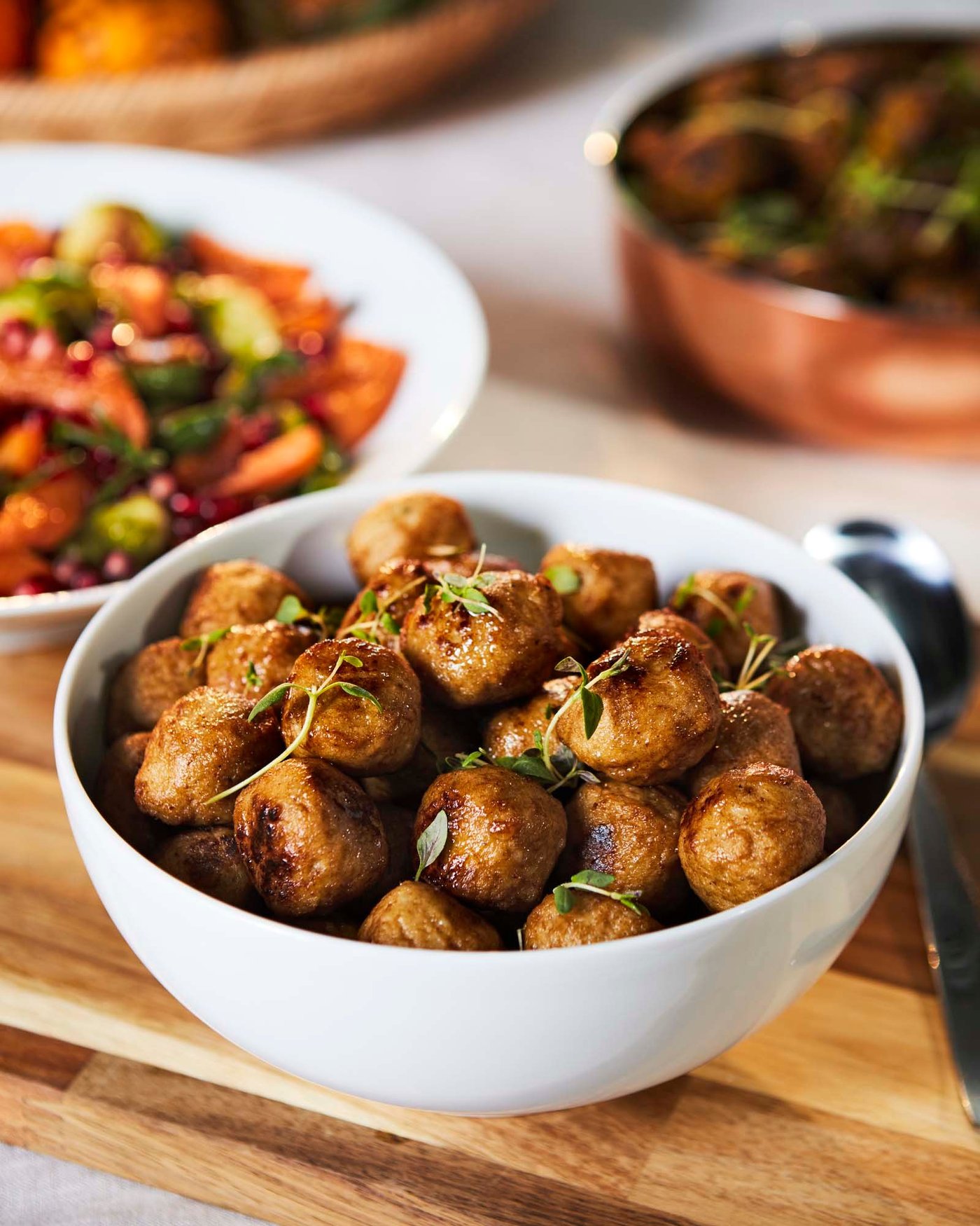 A bowl of IKEA chicken balls, garnished with herbs sit in a white bowl, on a wooden board. Behind are additional plates of food and a spoon.