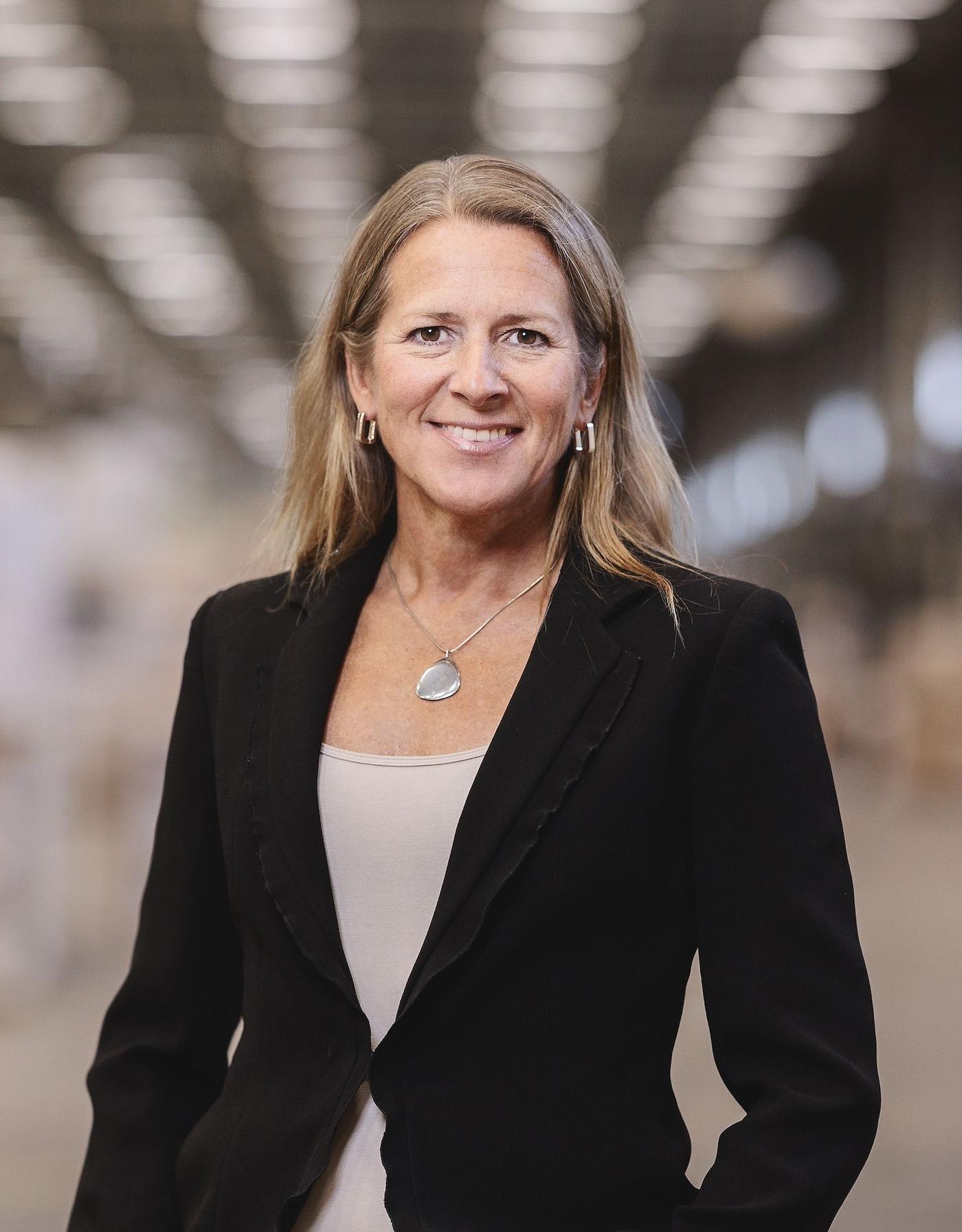 Portrait of Christina Niemelä Ström, who is wearing a black blazer with a blurred industrial space in the background.