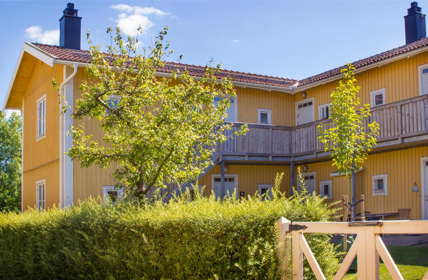 The facade of a yellow, wood BoKlok apartment building, with bushes and trees in the foreground on a sunny day.