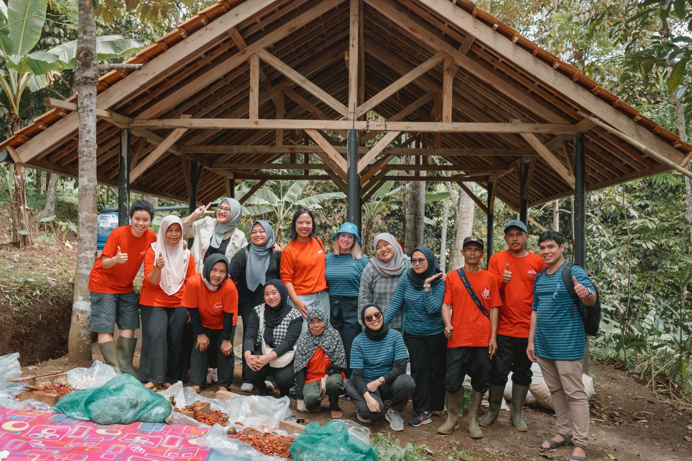 A group of people wearing matching red and blue shirts posing together under a wooden open-frame shelter in a forested area, with bags of agricultural produce placed on the ground in front of them.