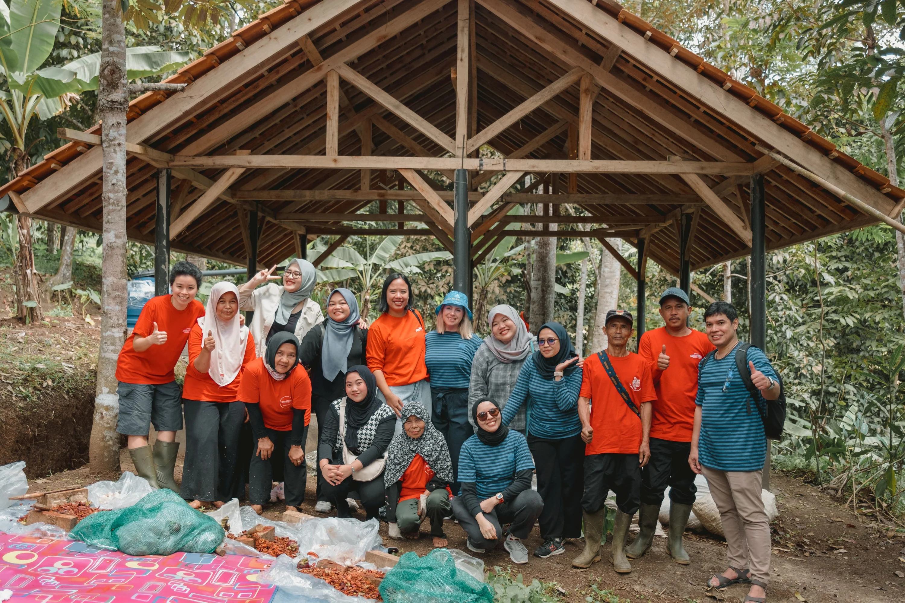 A group of people wearing matching red and blue shirts posing together under a wooden open-frame shelter in a forested area, with bags of agricultural produce placed on the ground in front of them.
