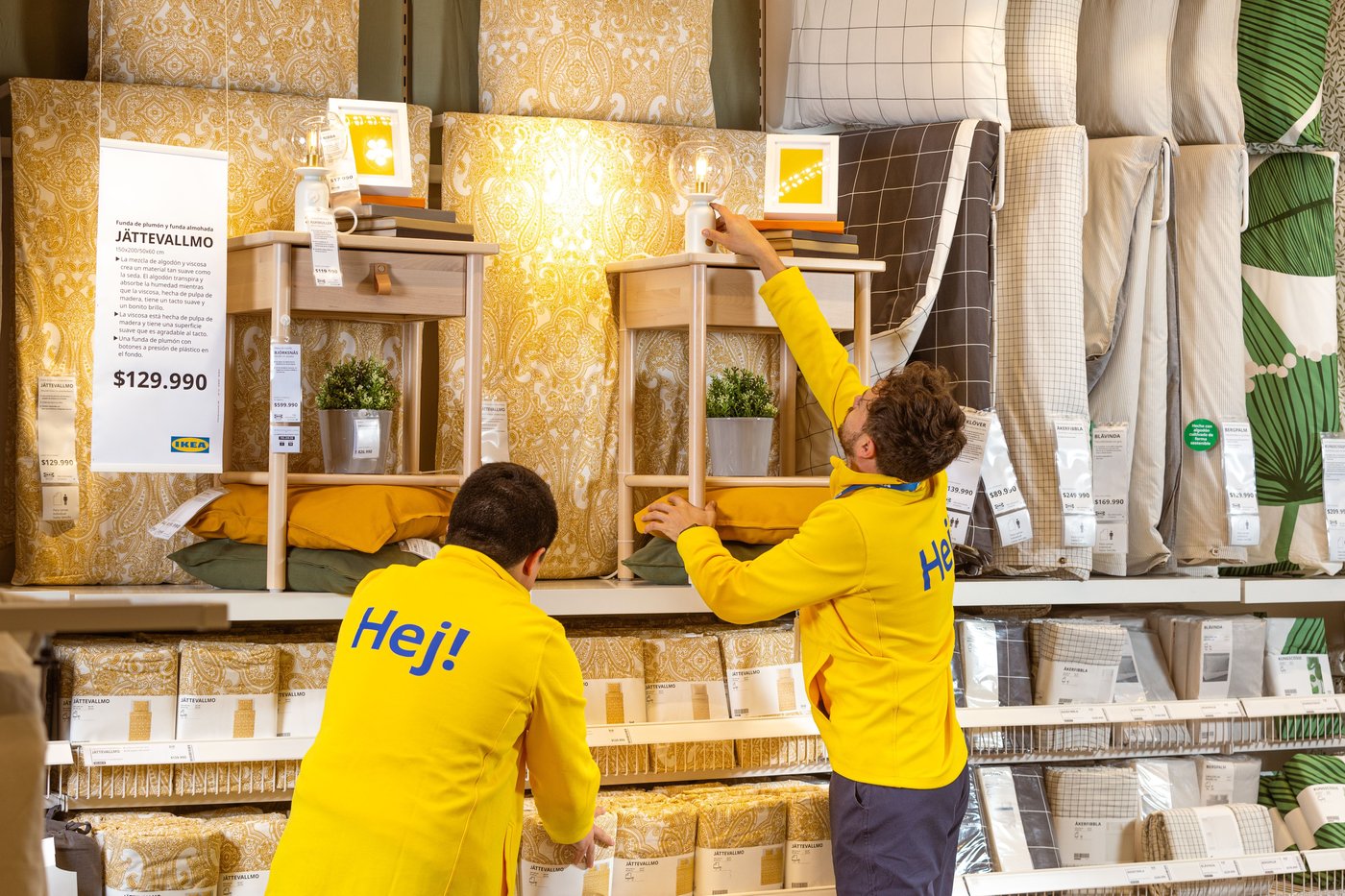 A shopping wall filled with bedcovers and night stands. Meanwhile two co-workers are working on making everything look neat.
