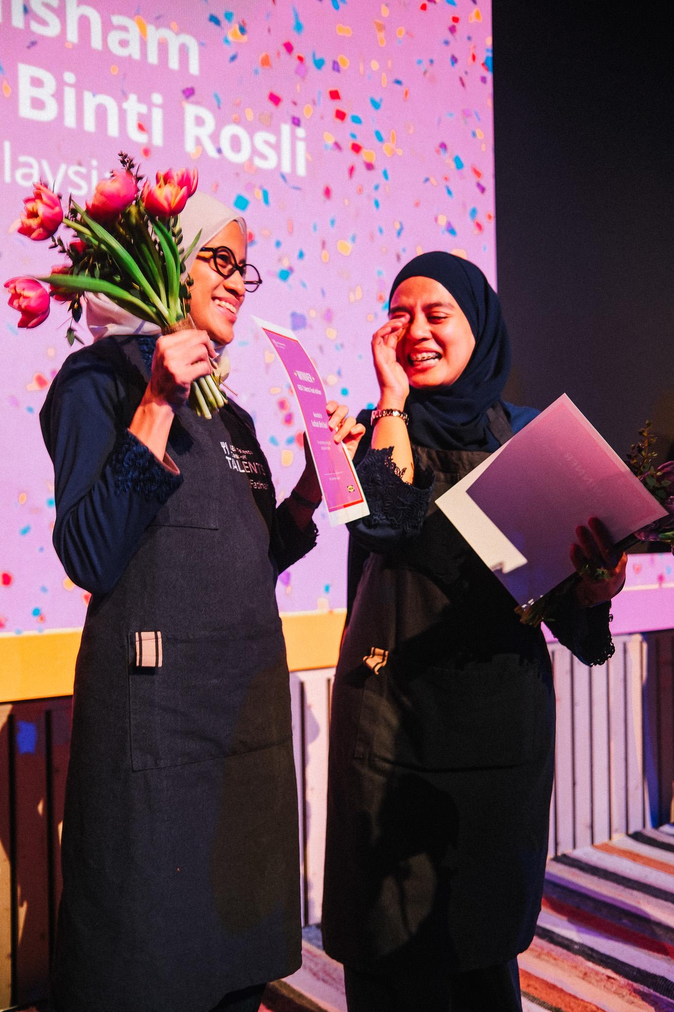 Two people with a bouquet of flower and open book, against a festive backdrop decorated with colourful dots and visible text. Both are wearing dark aprons, creating a celebratory atmosphere.