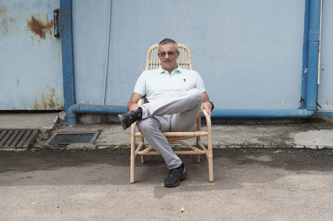 A person sits with legs crossed on a SALNÖ rattan armchair, outside a blue industrial building.