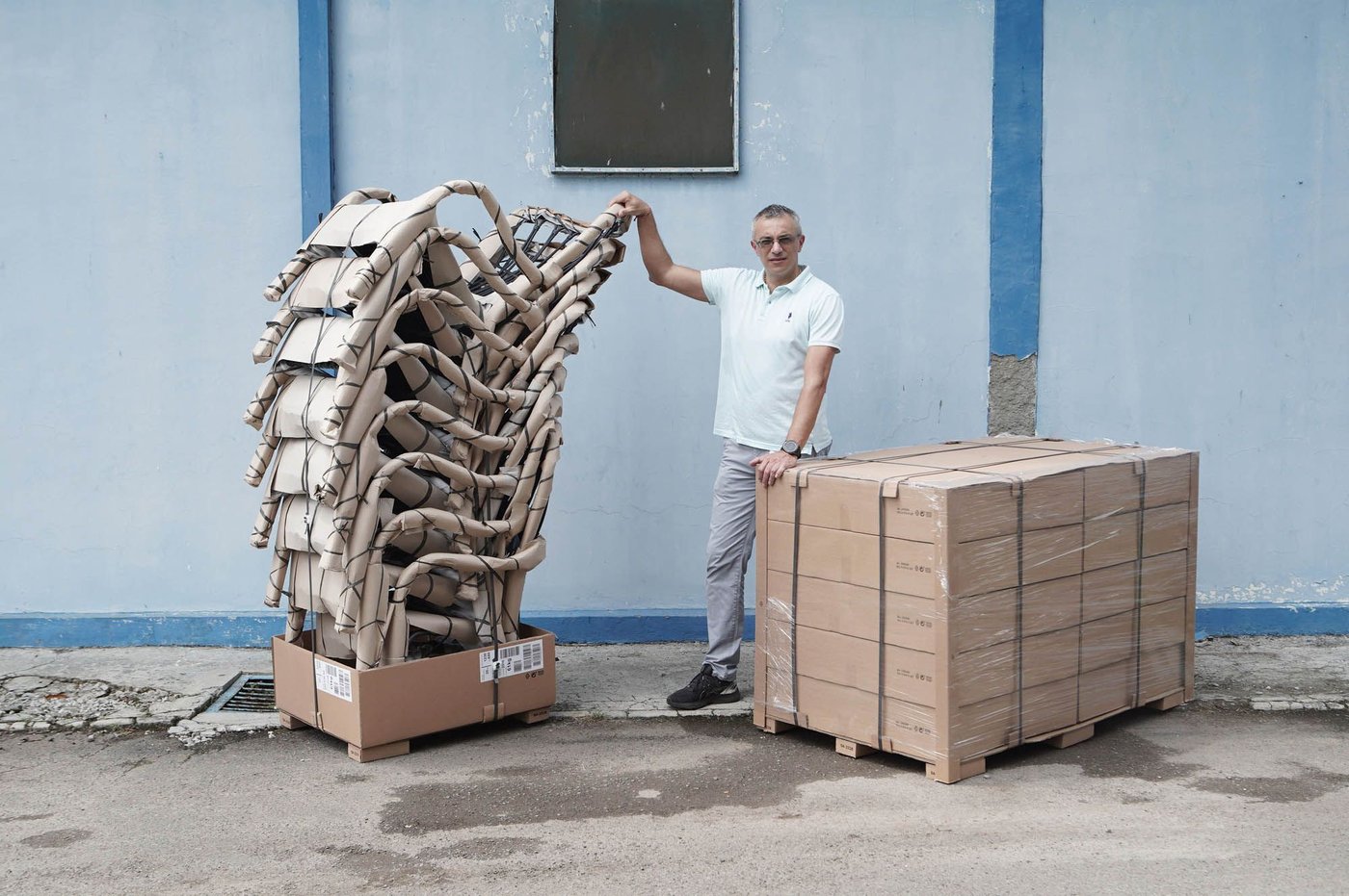A person stands in front of a light blue industrial building, with one hand reaching out to touch a tall stack of SALNÖ armchairs.