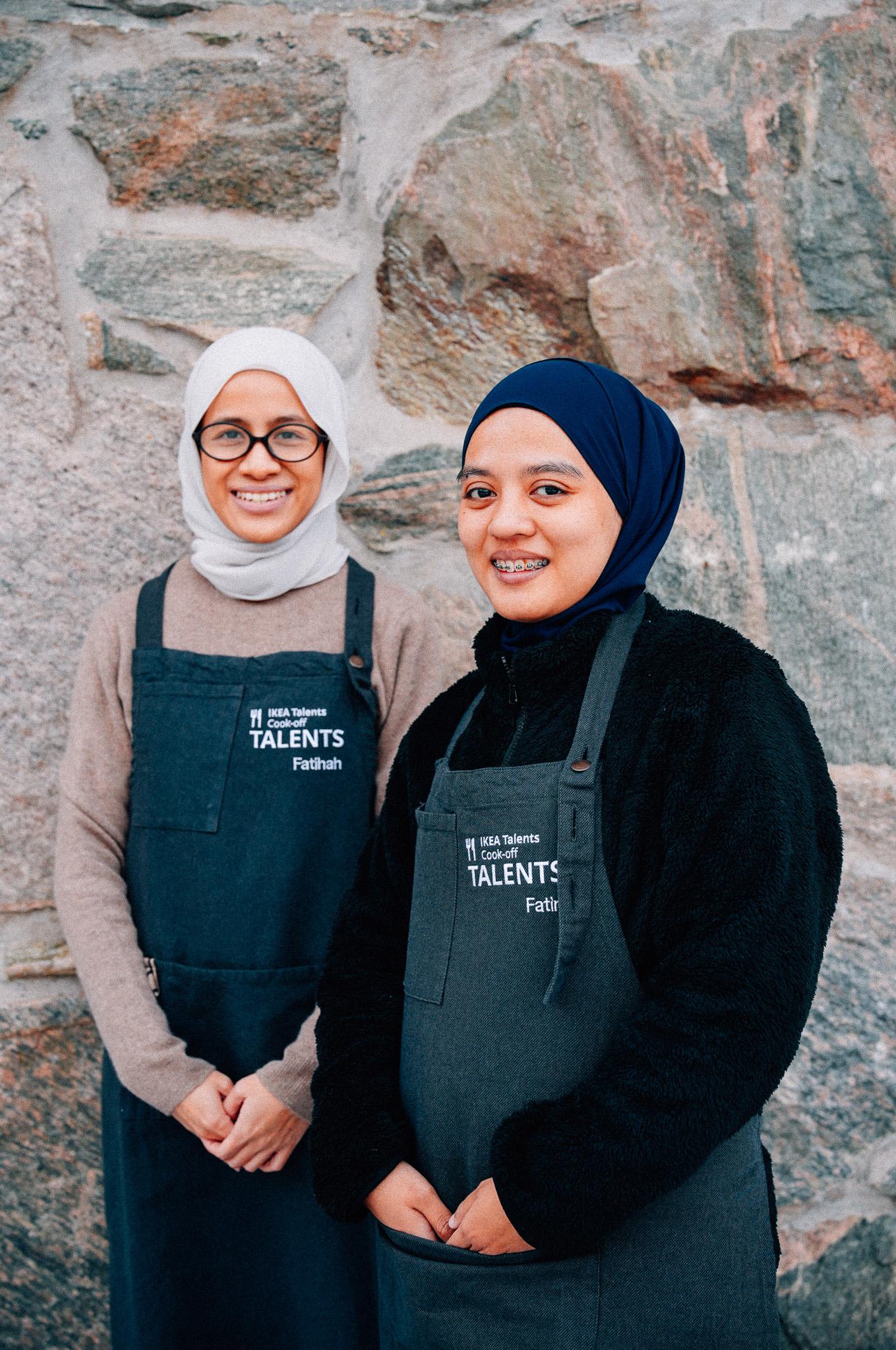 Two people wearing dark aprons over sweaters, standing in front of a textured stone wall. The apron features embroidered text, that reads "IKEA Talents Cook-off.