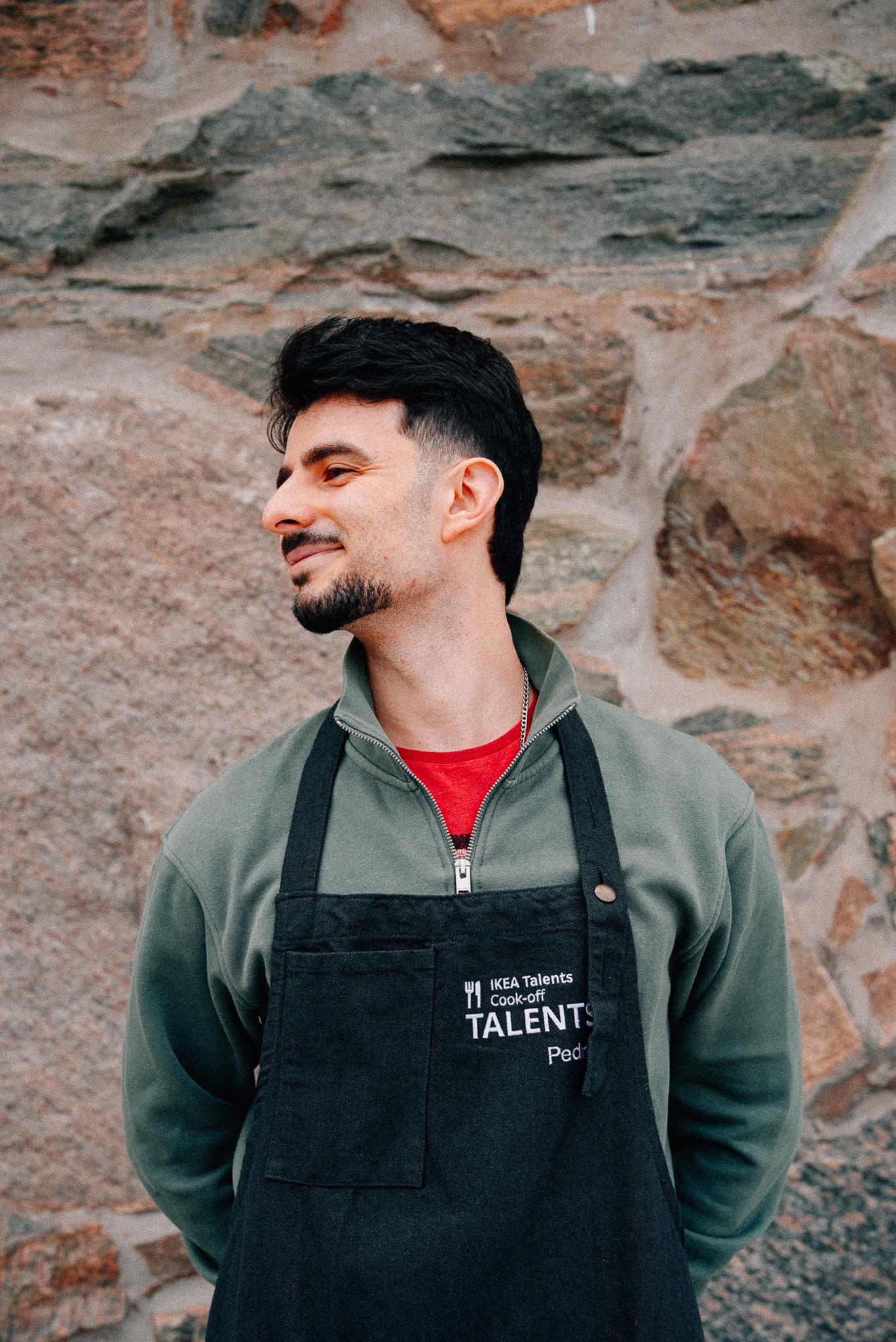A person wearing a dark apron over a sweater stands in front of a textured stone wall. The apron features embroidered text, that reads "IKEA Talents Cook-off.