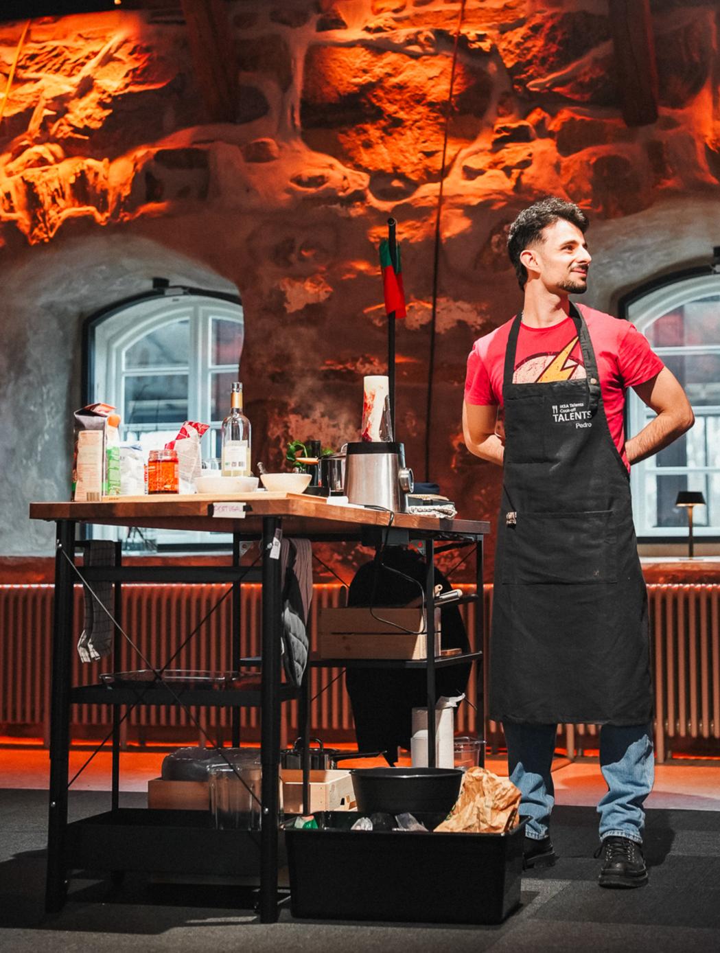 A person in a red shirt and apron stands next to a kitchen workstation filled with cooking tools and ingredients. The room features rustic stone walls and arched windows, creating a warm ambiance.