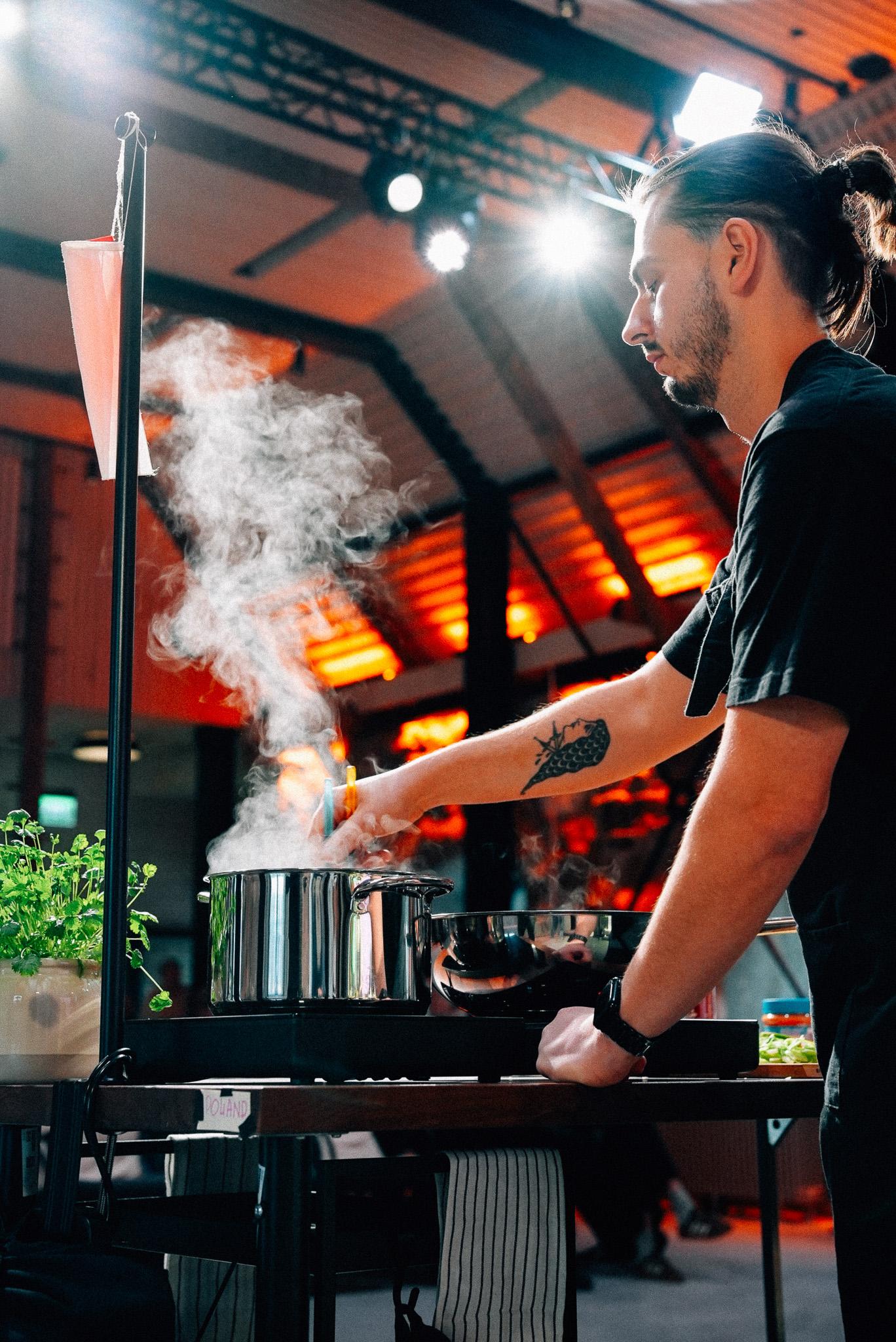 A person in a kitchen setting tends to a pot emitting steam, with warm lighting and a modern interior. The scene includes cooking ingredients and tools, creating a dynamic culinary atmosphere.
