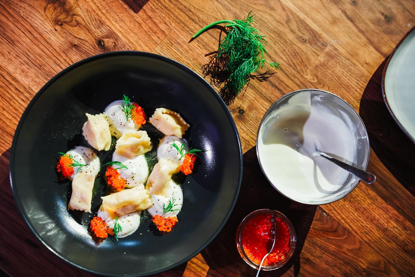 A dark bowl on a wooden table holds a dish with a small portion of pasta, dollops of cream sauce, and vibrant red garnishes, topped with herbs. Nearby are bowls of white and red condiments, adding to the colourful presentation.