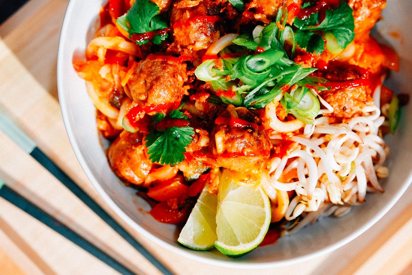 A bowl of vibrant noodle dish with meatballs, garnished with fresh herbs, sliced green onions, and lime wedges. The presentation includes bean sprouts, creating a colourful and appetising meal. Chopsticks rest on the side.